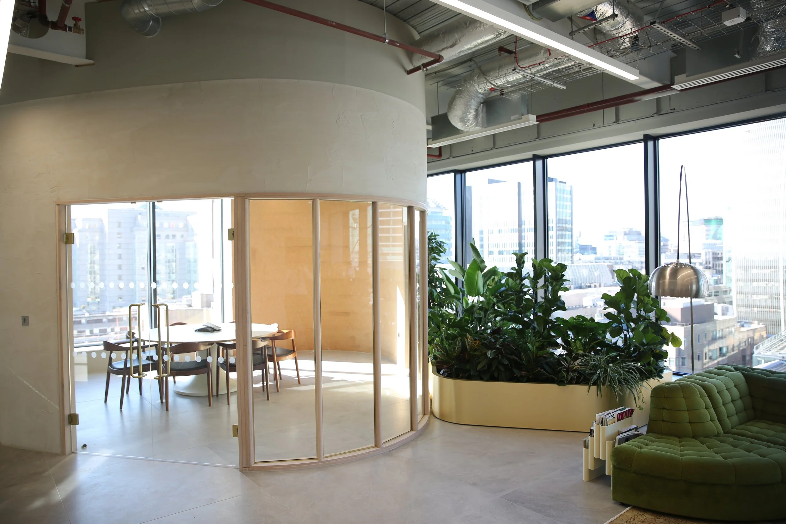 Modern office space with large windows showing city skyline. Contains a circular planting area filled with greenery, a green tufted couch, a small magazine stand, and a wooden table with chairs in a glass-enclosed room.