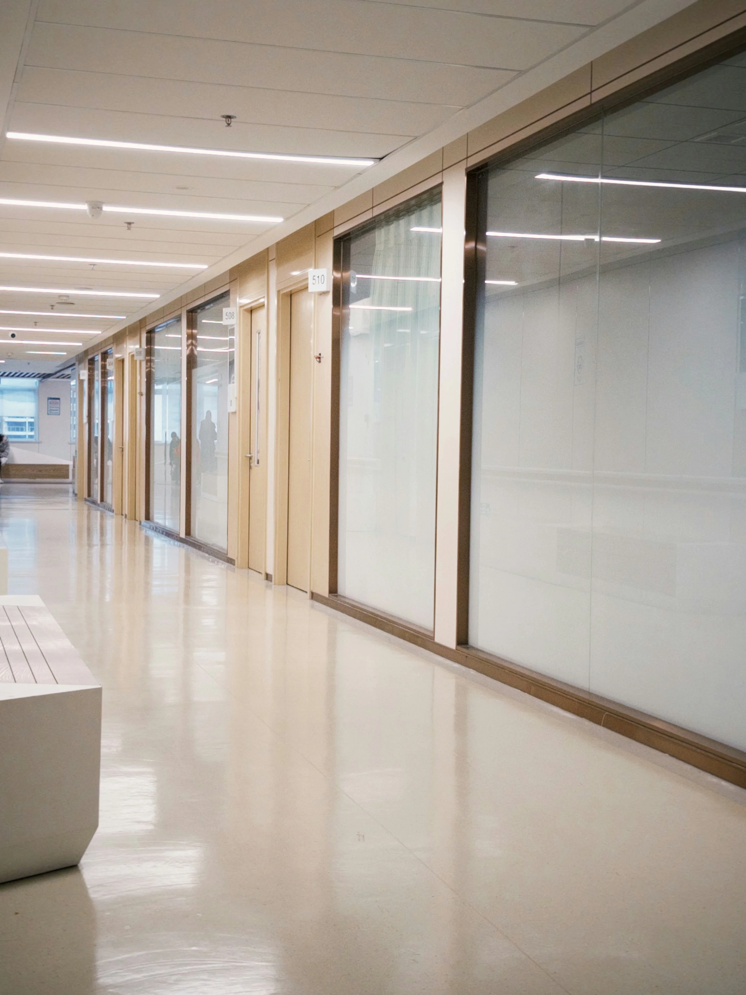Empty indoor corridor with glass doors, ceiling lights, and polished beige floor reflecting light, with a white bench and people visible in the background.