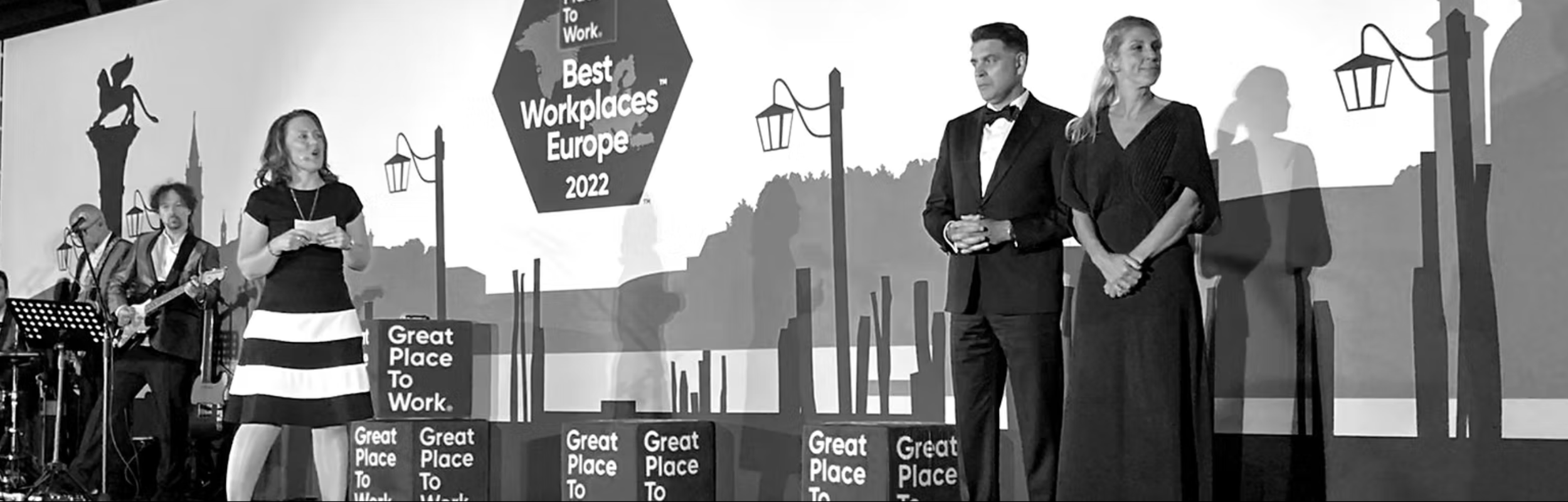People standing on stage at the Best Workplaces Europe 2022 event, with a woman speaking into a microphone and others listening, in front of a backdrop with company branding.