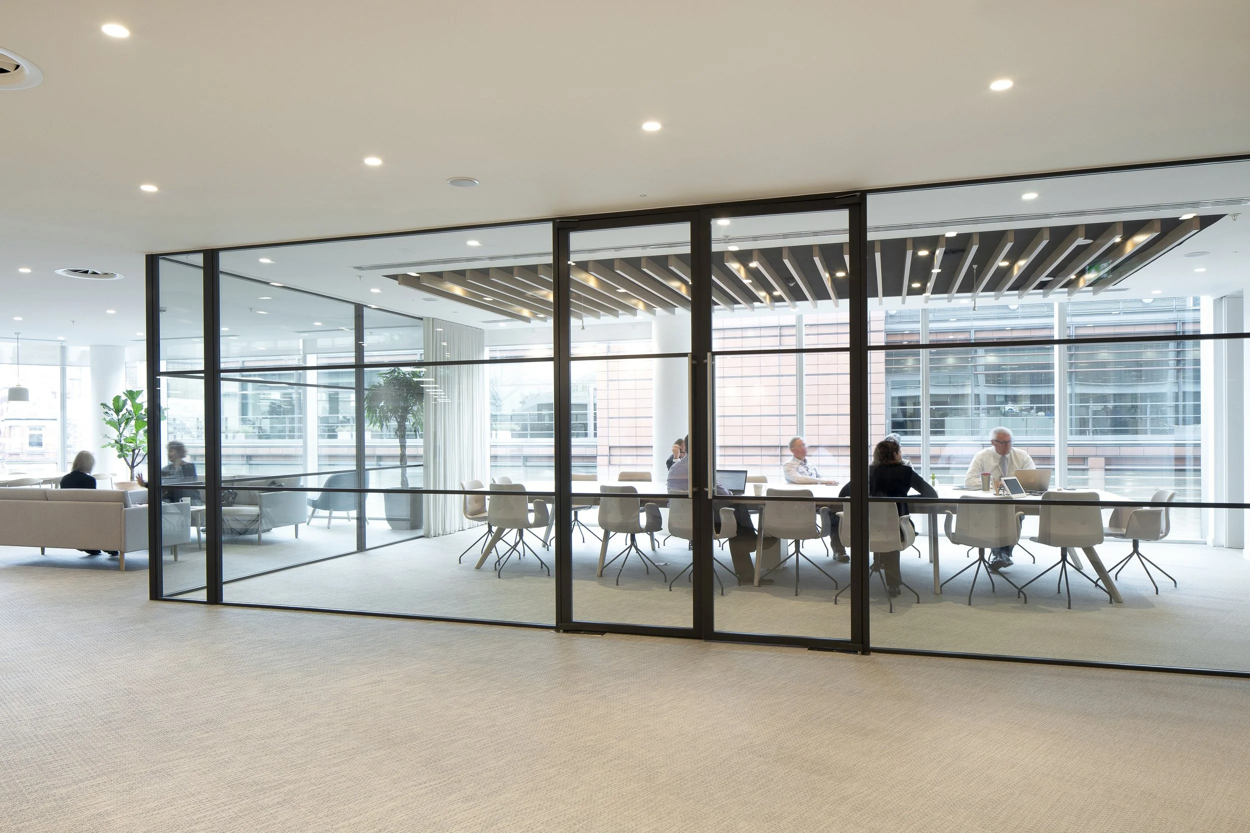 Modern conference room with glass walls, white chairs, and a group of people working on laptops and reading, with large windows outdoor city view.