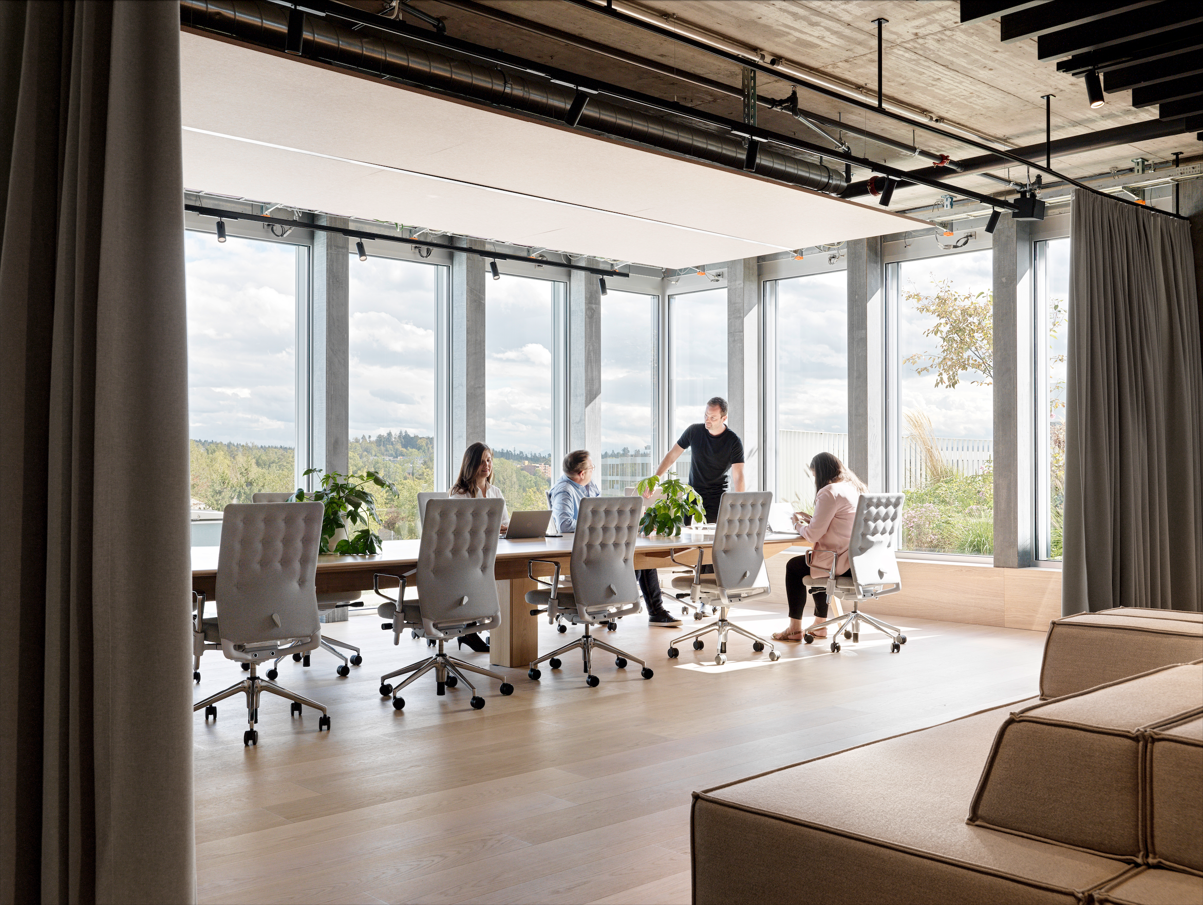 People sitting around a table in a modern office conference room with large windows, curtains, and ceiling industrial pipes.