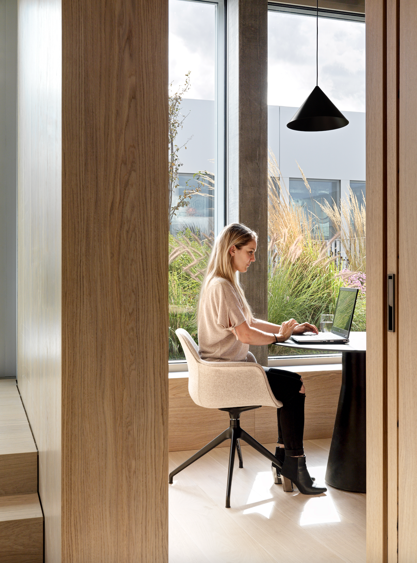 A woman working on a laptop at a small round table in a room with large windows and modern decor, surrounded by wooden walls and a view of a garden outside.