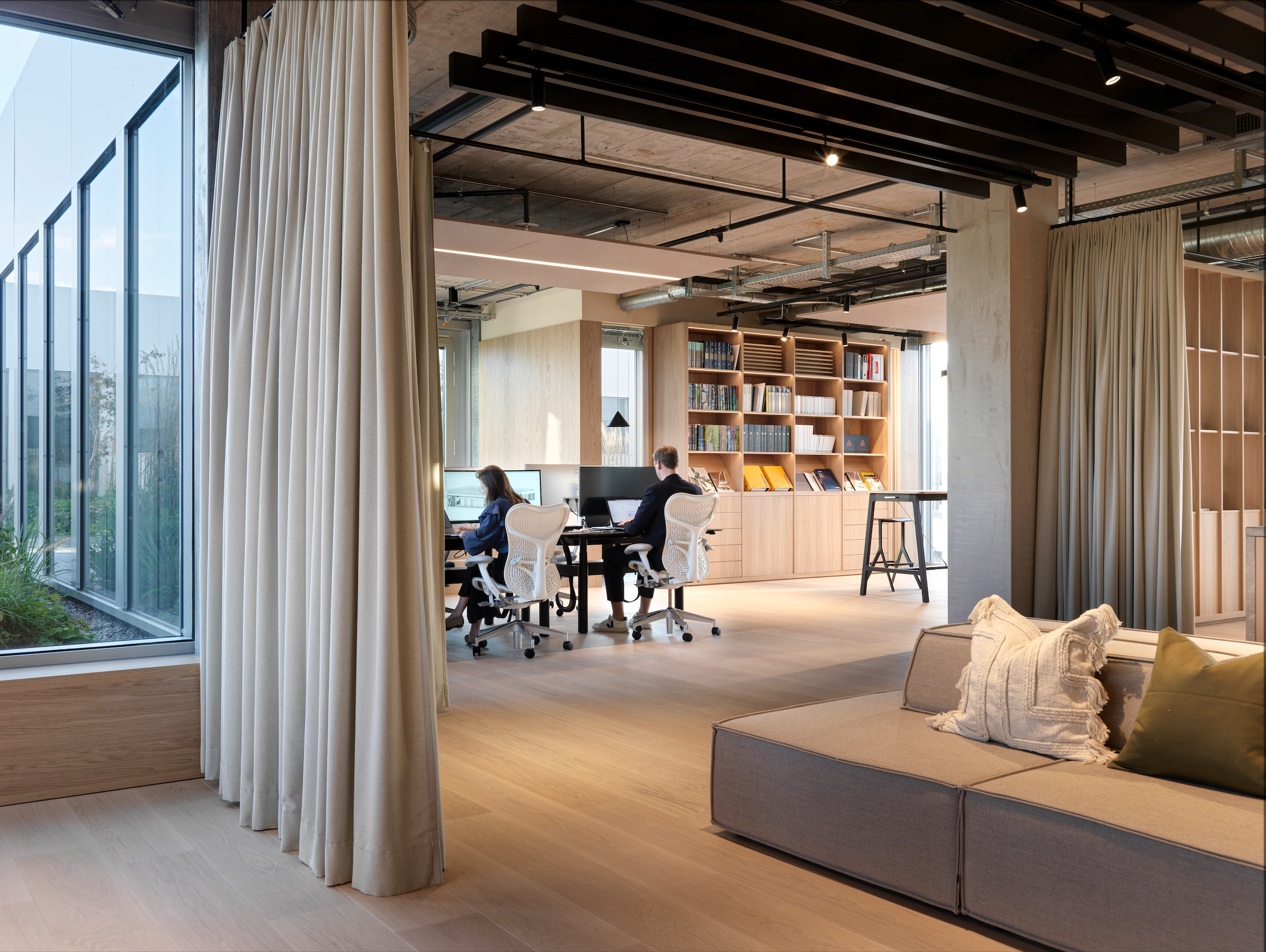 Modern open-plan office space with large glass windows, wooden bookshelves, and people working at desks. There is a cozy seating area with cushions in the foreground.