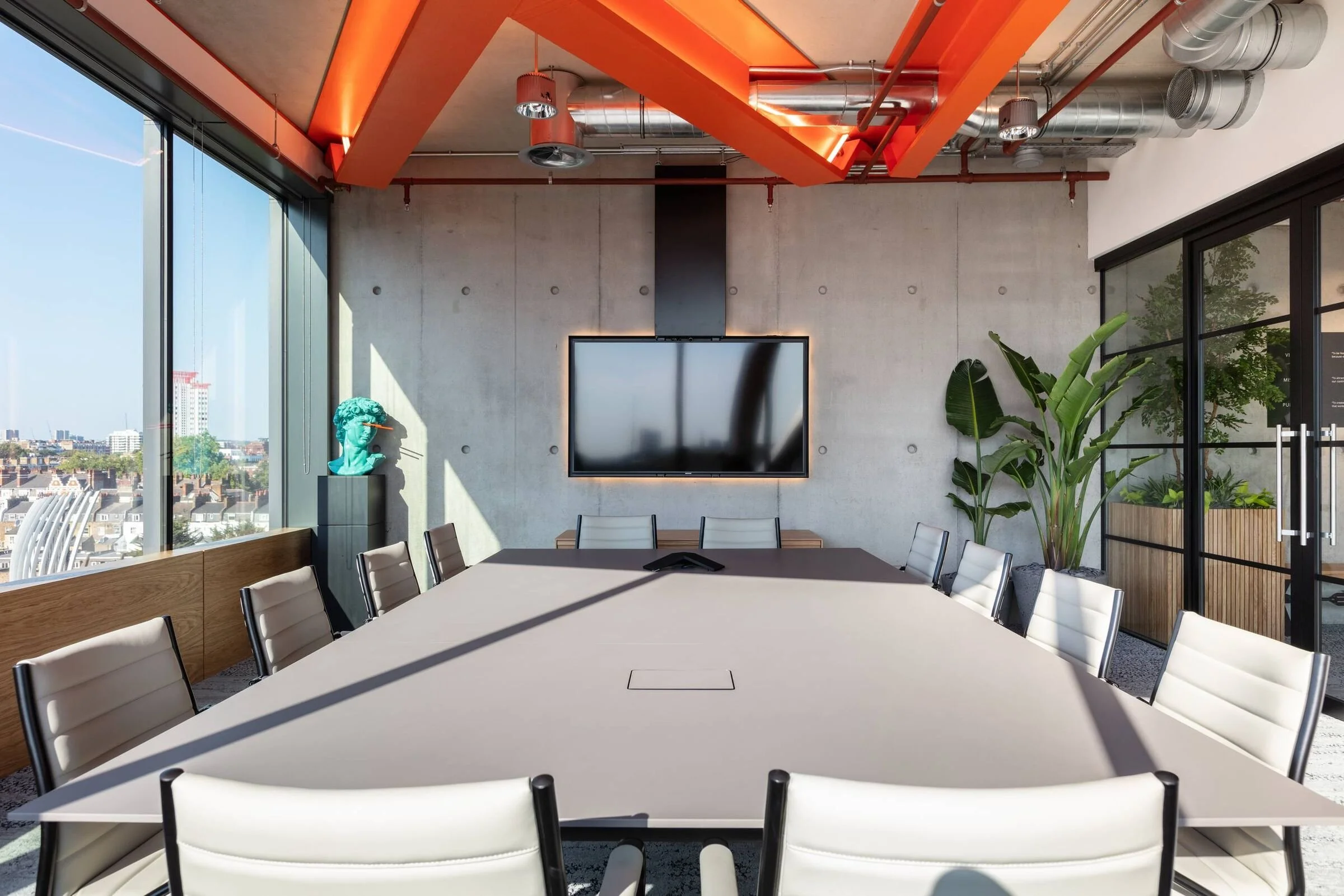 Modern conference room with a large table and white chairs, concrete wall with TV, large windows with city views, green plants, and exposed ductwork and orange structural elements on the ceiling.