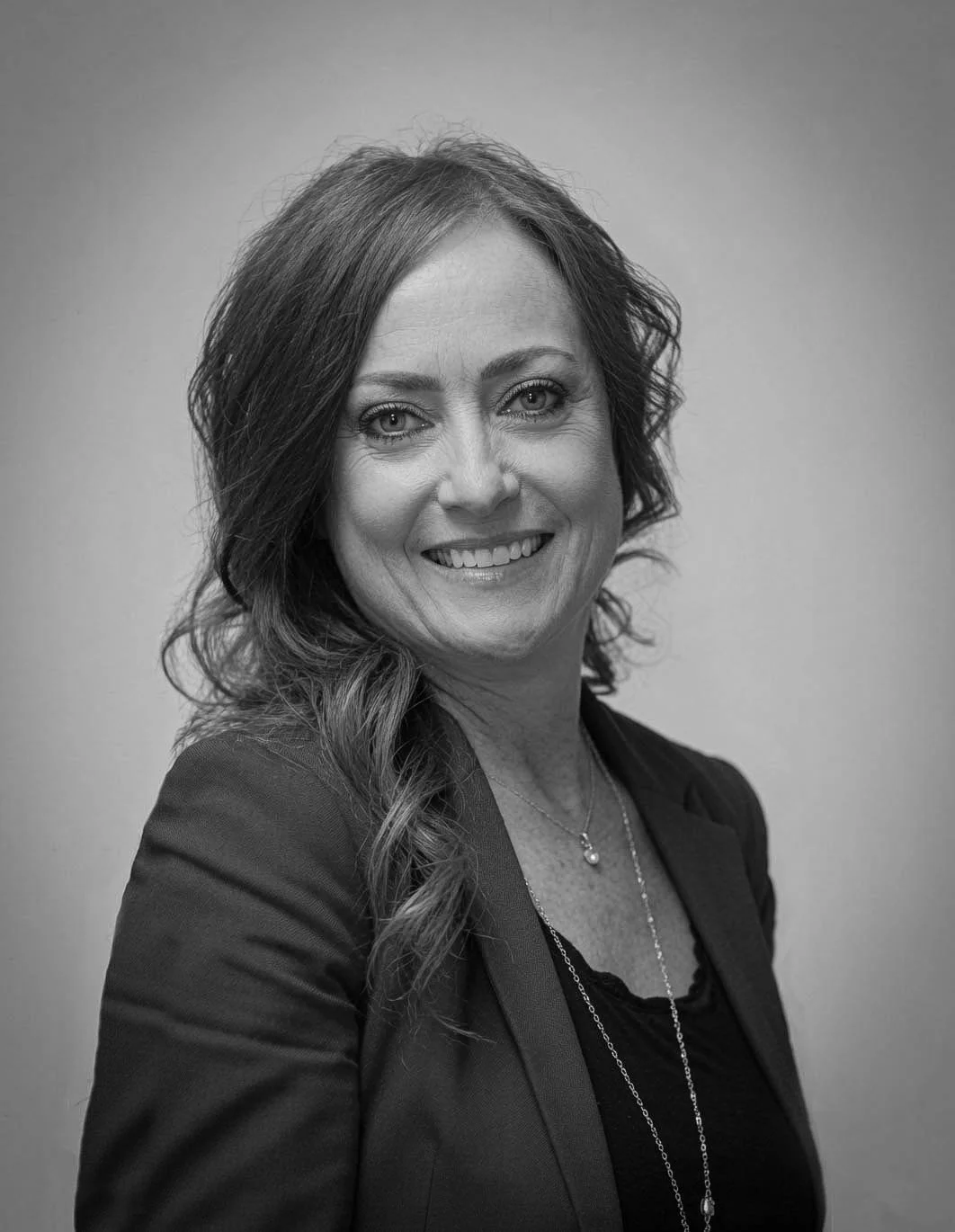 Black and white portrait of a woman with wavy hair smiling at the camera, wearing a blazer and a necklace.
