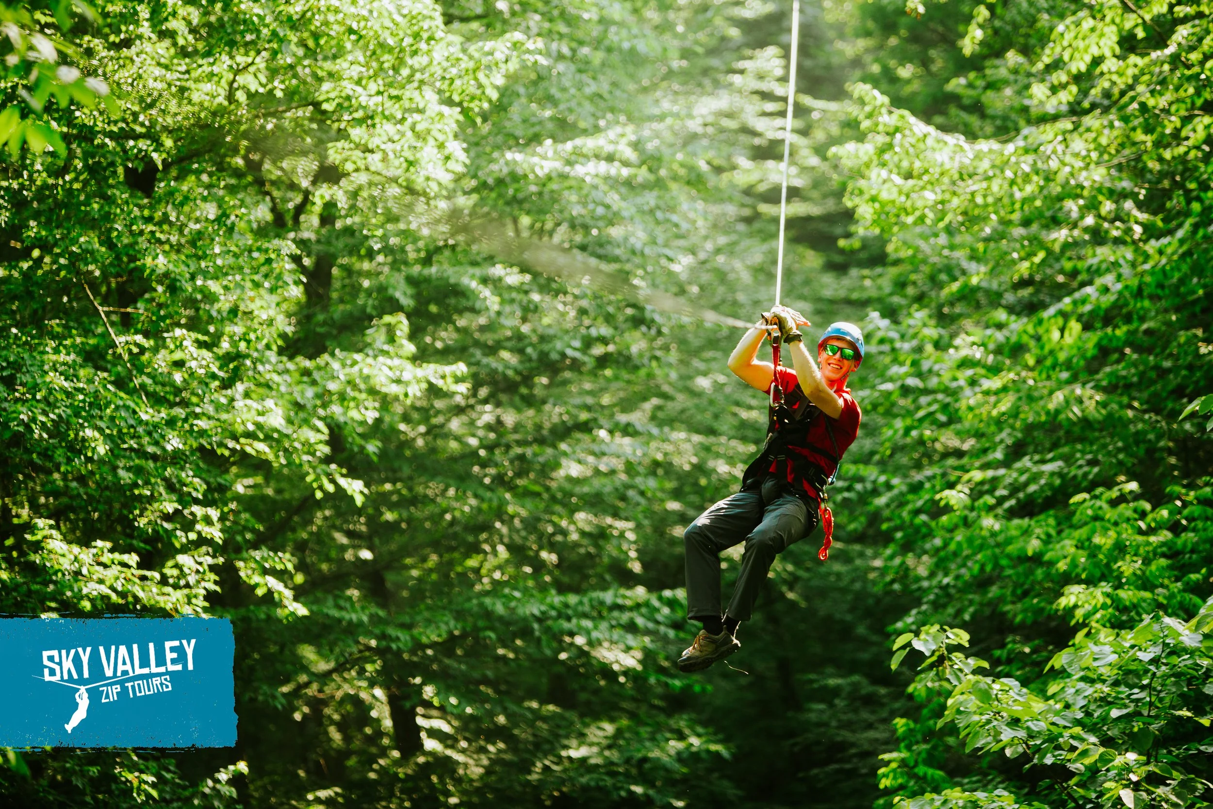 A person zip lining through a lush green forest, wearing a helmet and sunglasses, smiling, with a sign that says "Sky Valley Zip Tours" in the bottom left corner.