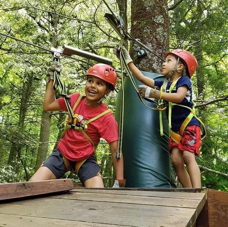 Two children wearing helmets and harnesses are on a zip line in a forest. One child is smiling and holding onto the zip line, while the other is reaching up towards the zip line cable.
