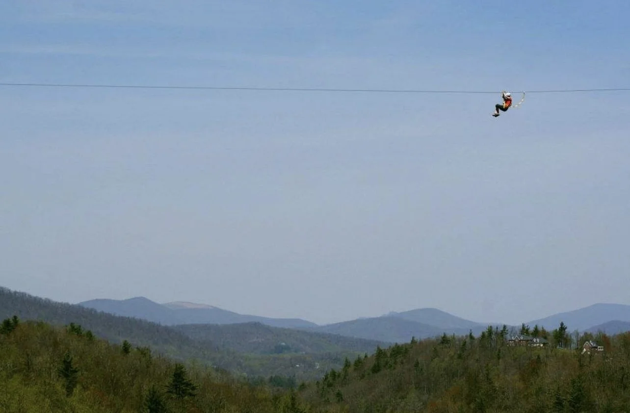 A person on a zip line flying over a scenic mountainous landscape with trees and hills.