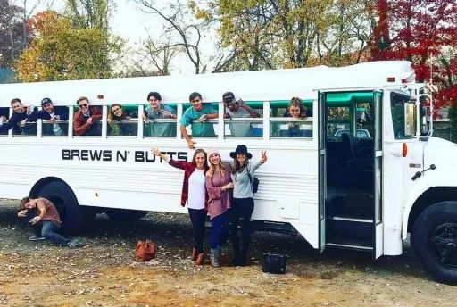 Group of people posing in and around a white beer bus in a scenic outdoor area during autumn.