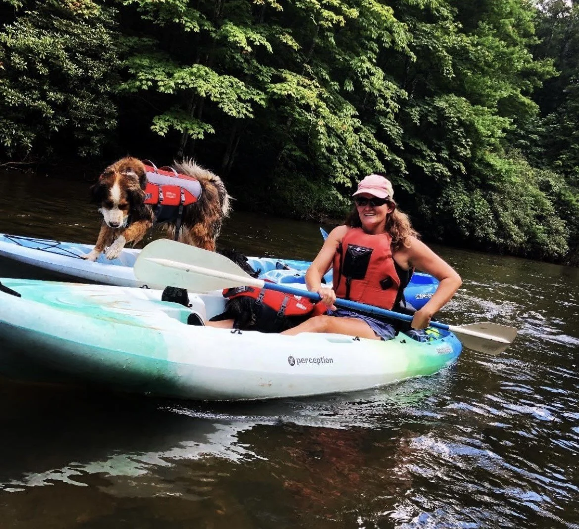 Woman and dog kayaking on a river surrounded by lush green trees.