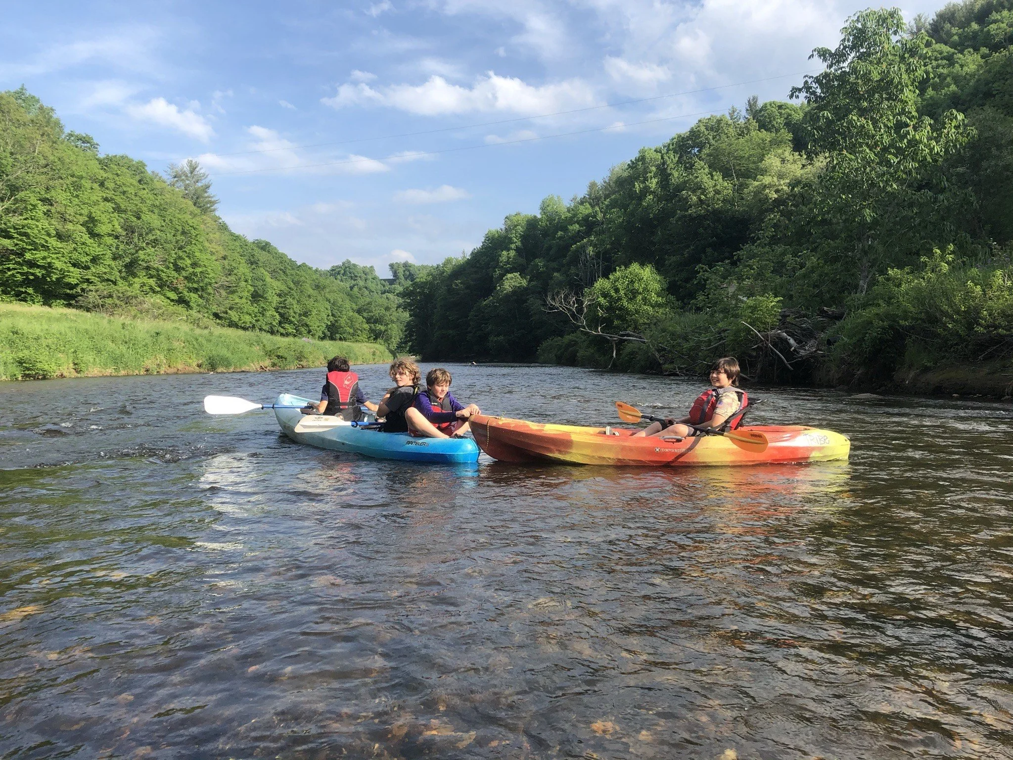 Four kids kayaking on a river surrounded by lush green trees, with a partly cloudy sky overhead.