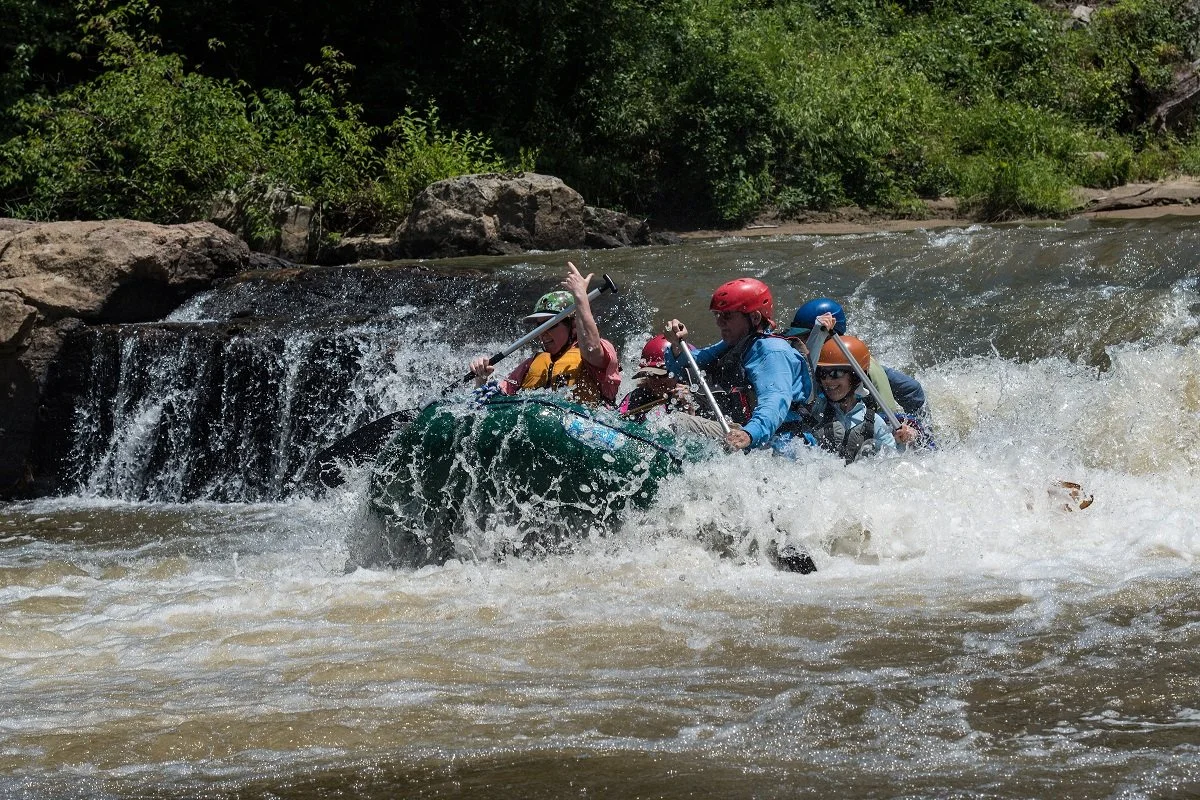A group of people white-water rafting on a river with a small waterfall, wearing helmets and life jackets.