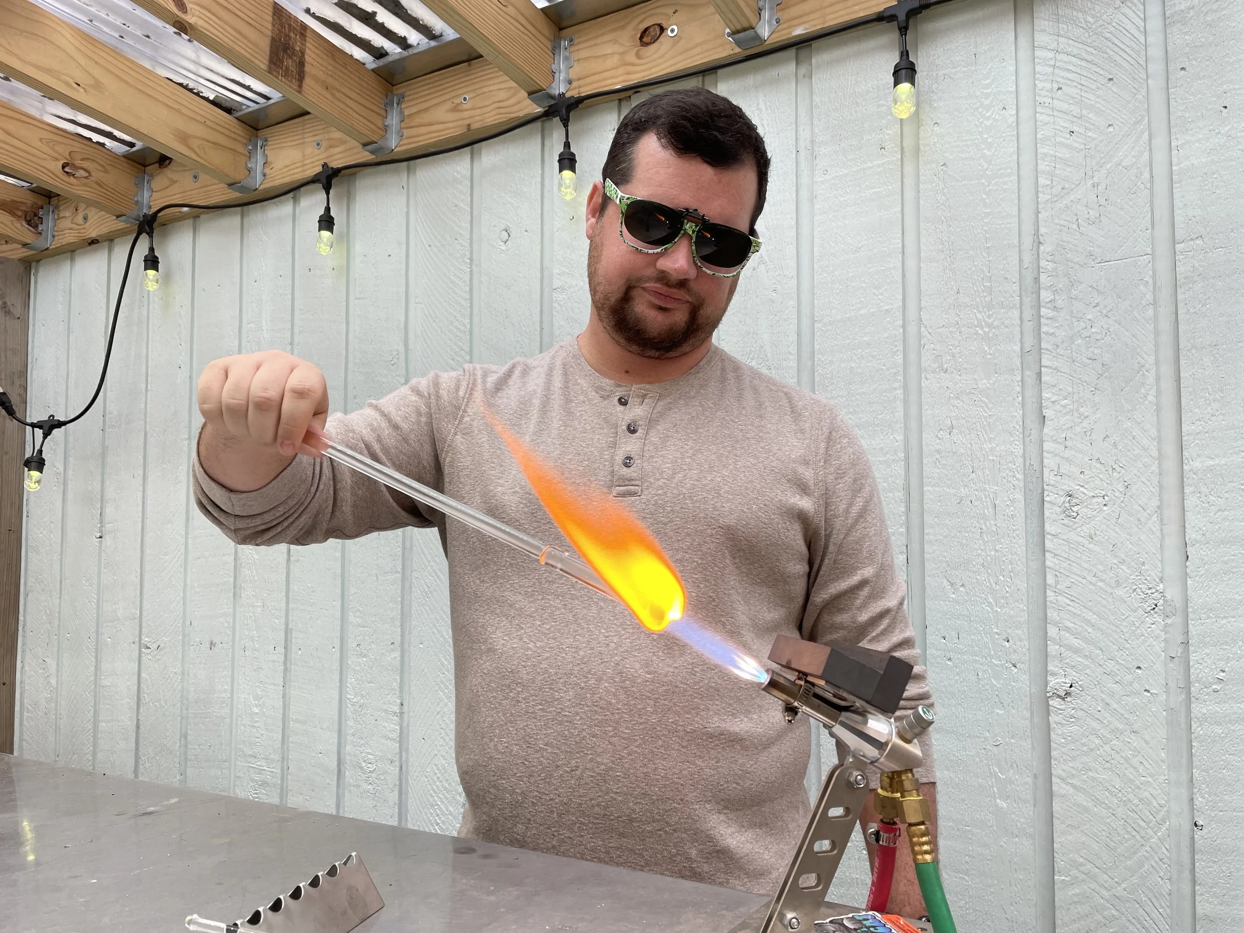 A man wearing sunglasses and a beige long-sleeve shirt is holding a glass tube in a flame produced by a propane torch, with a clothespin holding the tube. He is working outdoors near a light-colored wooden wall with string lights overhead.