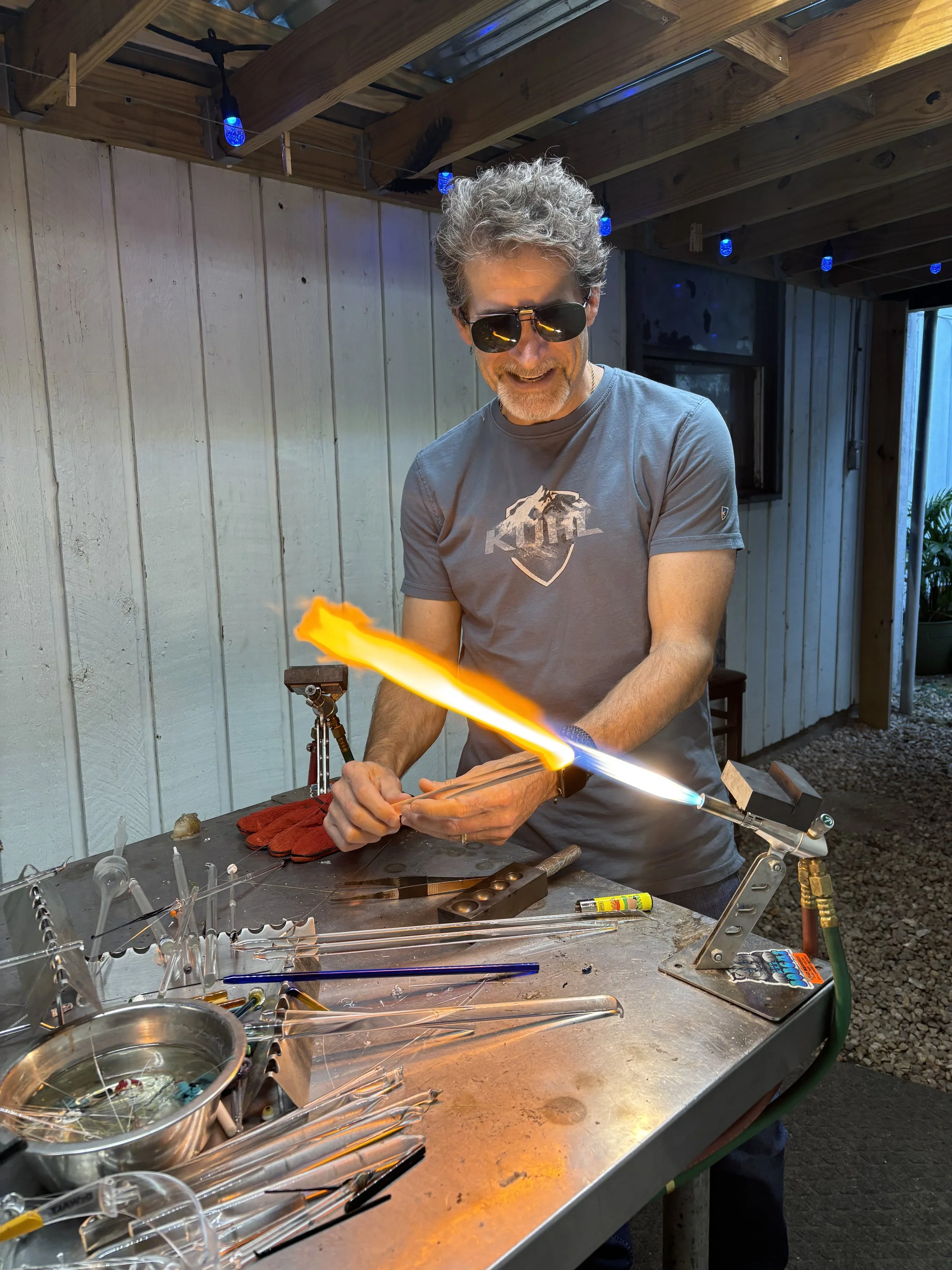 A man with sunglasses and a gray T-shirt is working in a glassblowing studio, using a torch to shape glass. The worktable in front of him holds various glassblowing tools, glass tubes, and a bowl of water. The background includes a wooden wall and ce