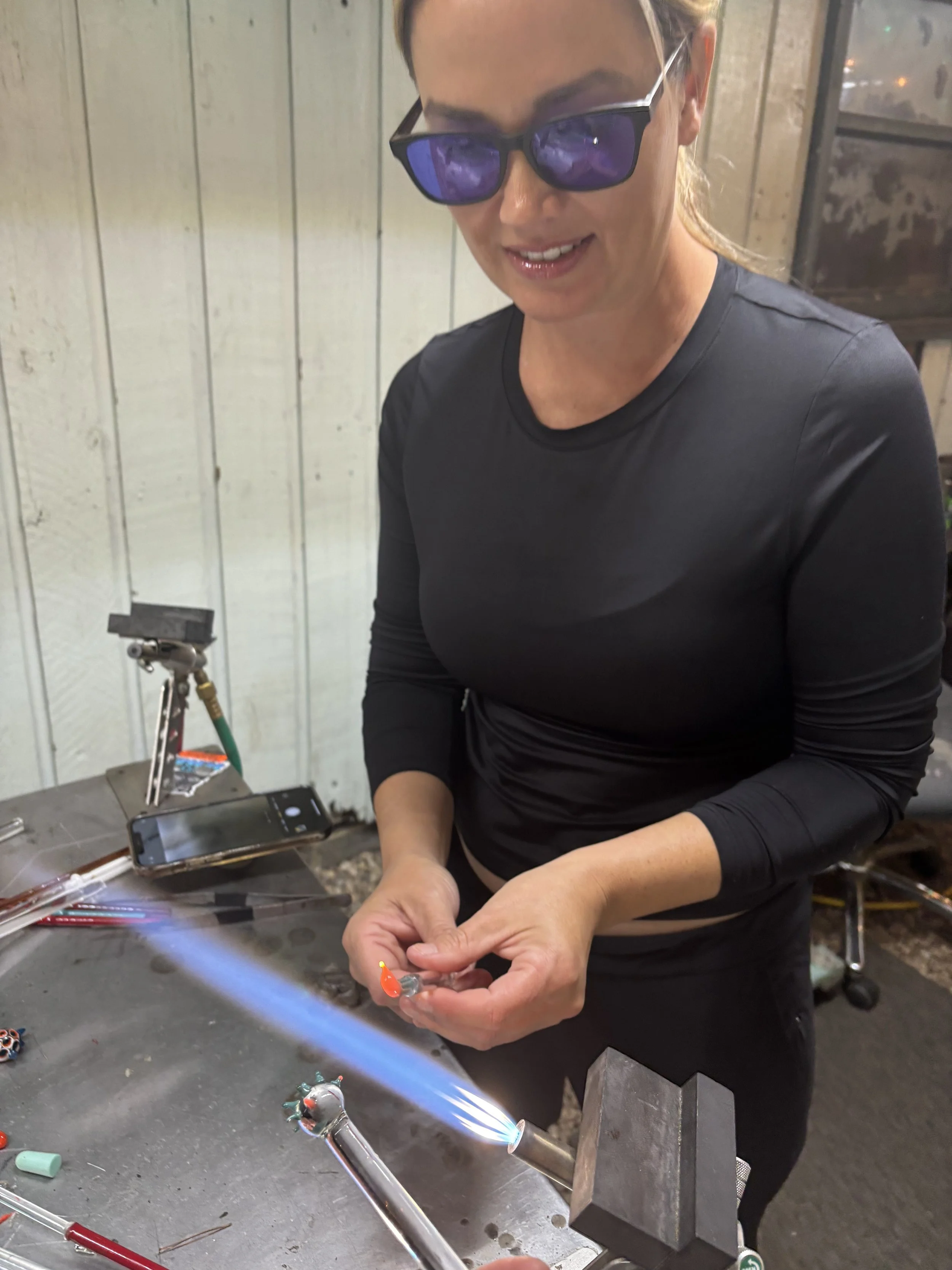 Woman working with glassblowing torch, wearing protective glasses, shaping glowing glass at a workshop.