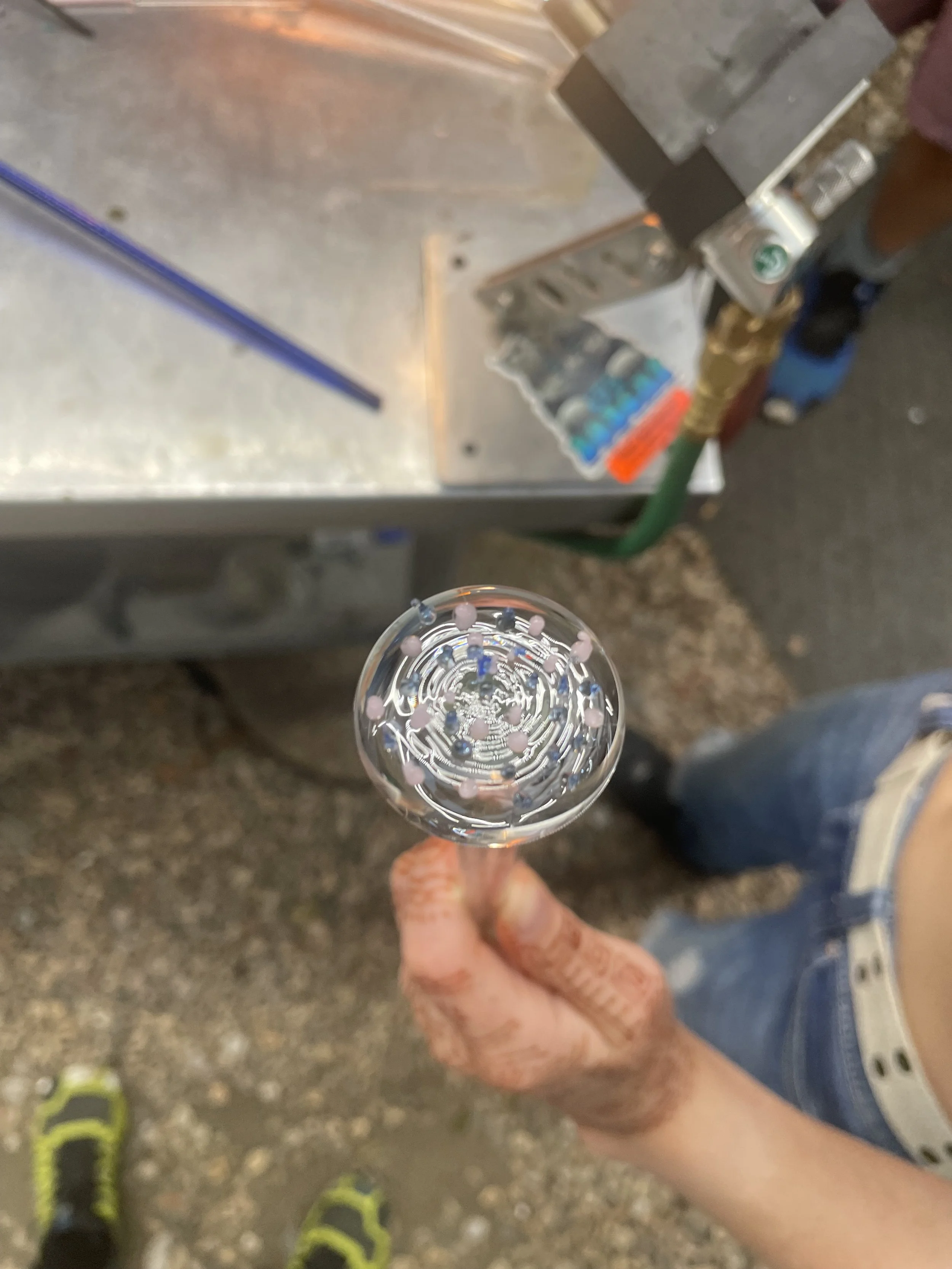 A person holding a glass of clear liquid with small colorful beads inside, with a metal workbench and tools in the background.
