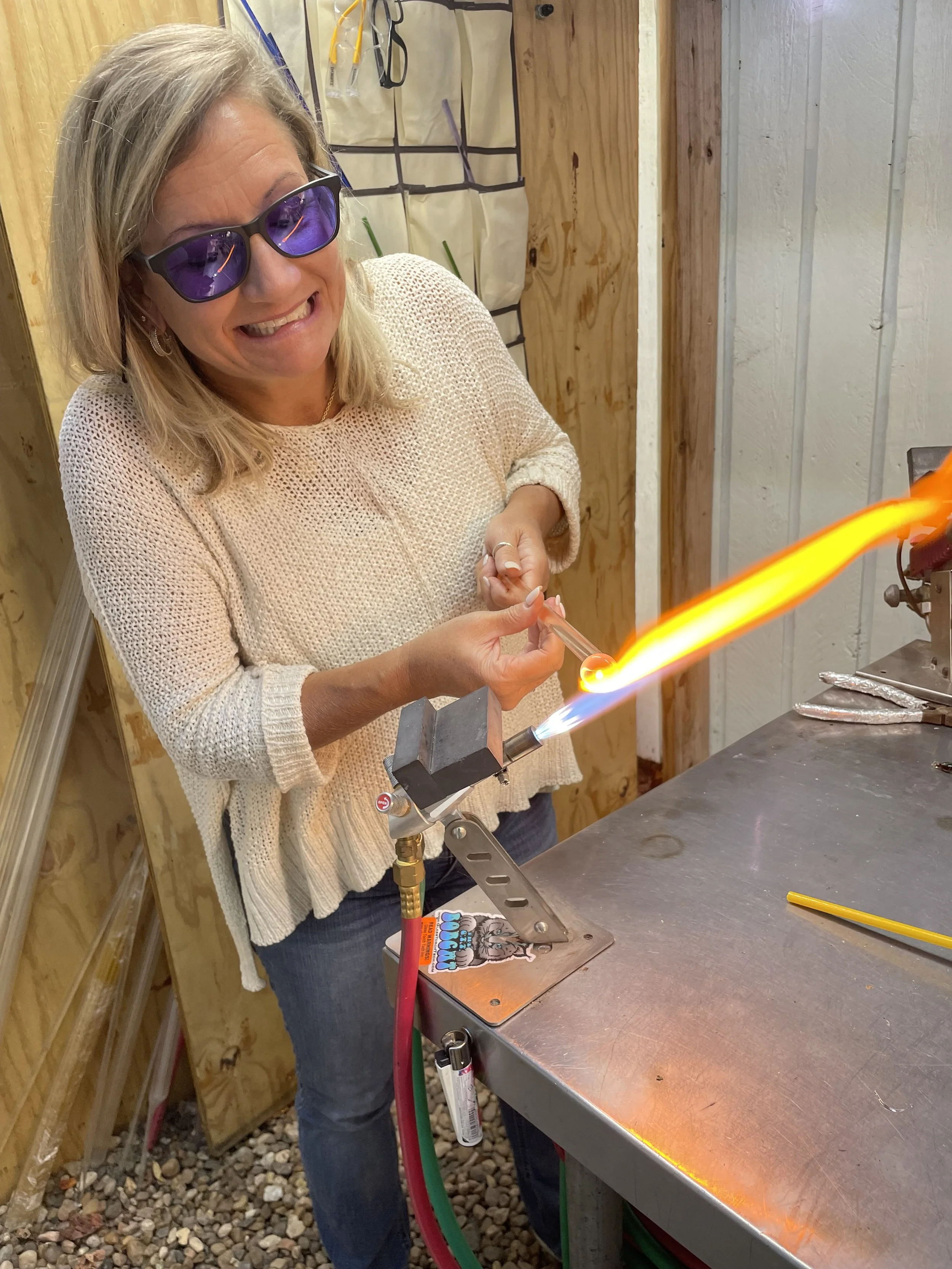 A woman wearing sunglasses and a beige sweater is working with a torch to heat or mold glass on a stainless steel worktable.