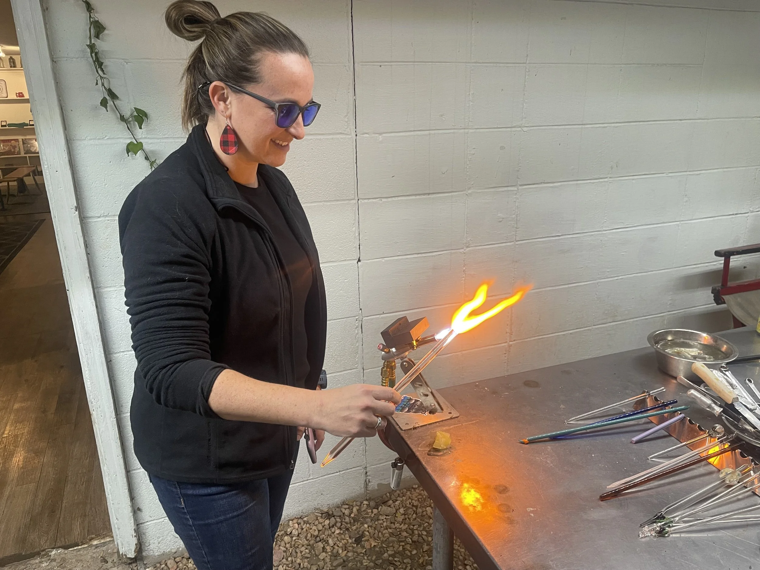 A woman wearing glasses and a black jacket is working with glass in a glassblowing workshop. She is holding a long glass tube that is emitting a flame, shaping it in the air. Various tools and glass pieces are on the metal worktable.