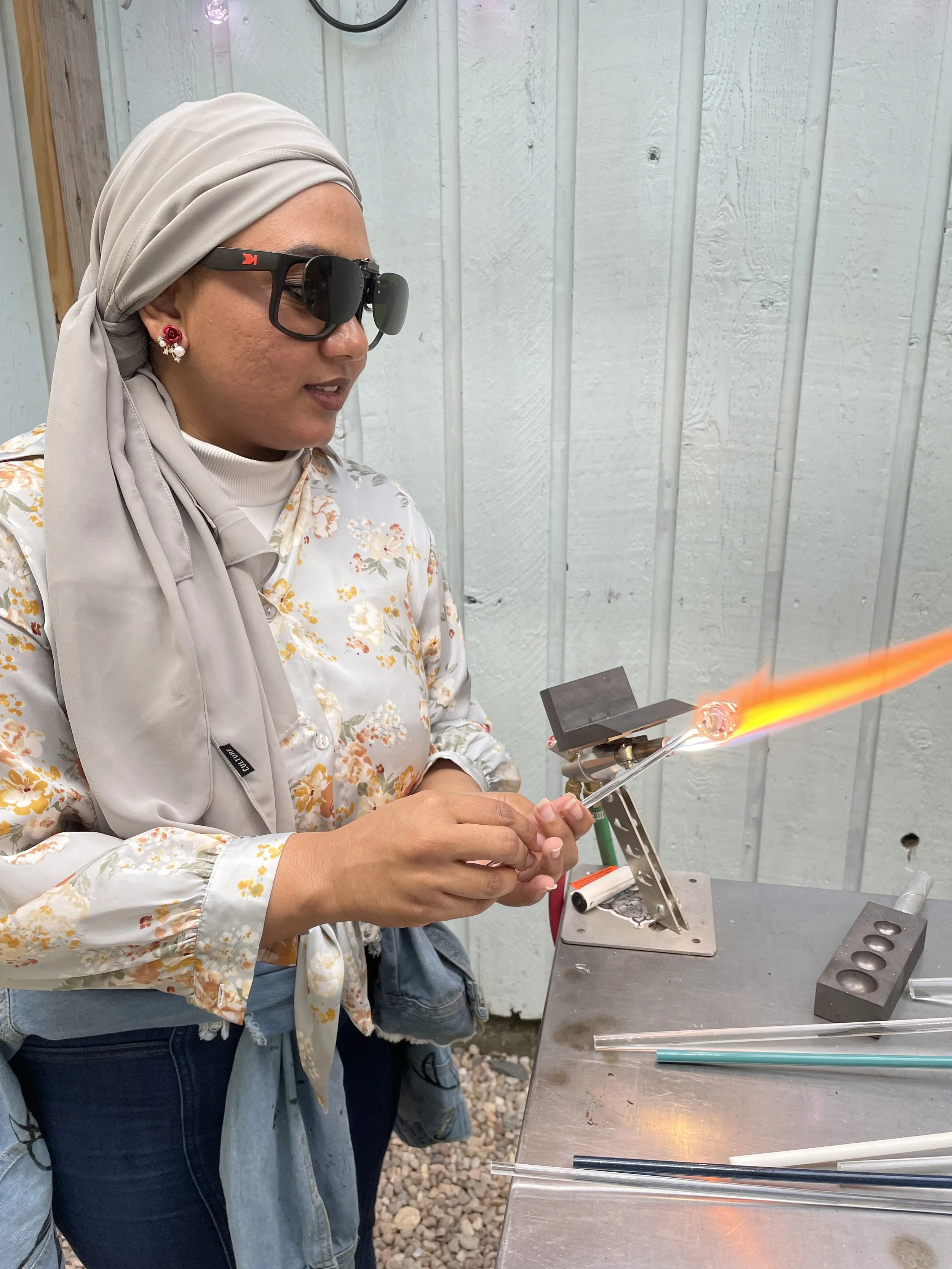 A woman wearing a headscarf, sunglasses, and floral blouse is working with glassblowing tools, shaping a glass piece with flame in a workshop setting.