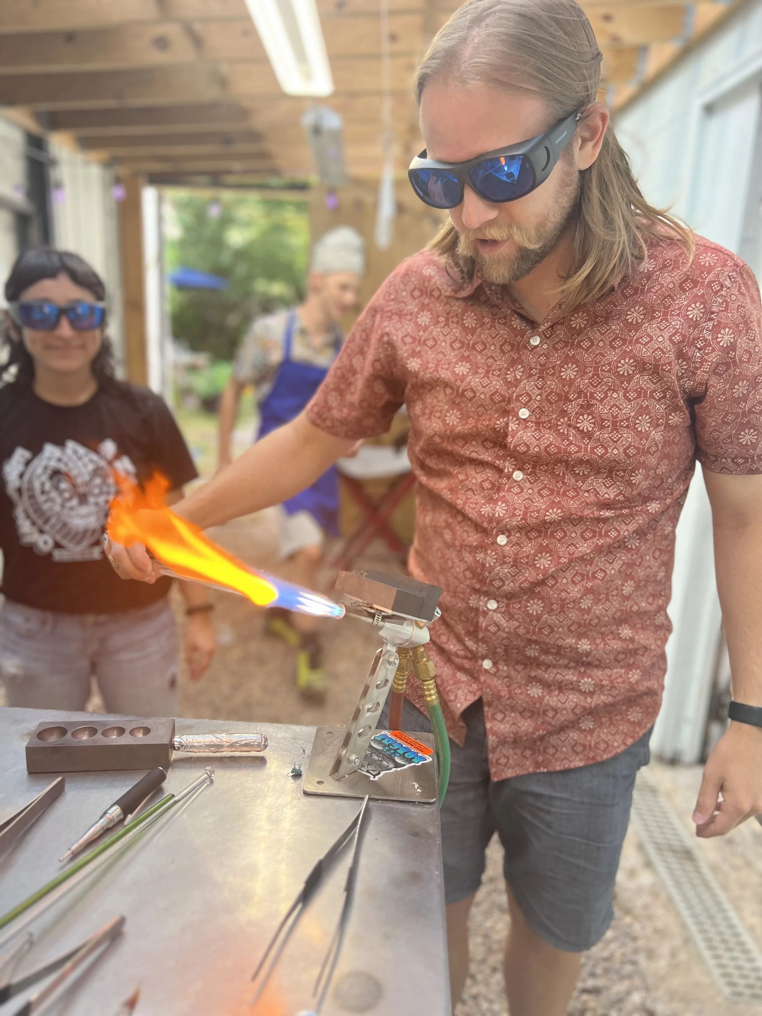 A man wearing safety glasses and a patterned red shirt is using a blowtorch to work on glass art, with two women in the background observing, in a workshop or outdoor workspace.
