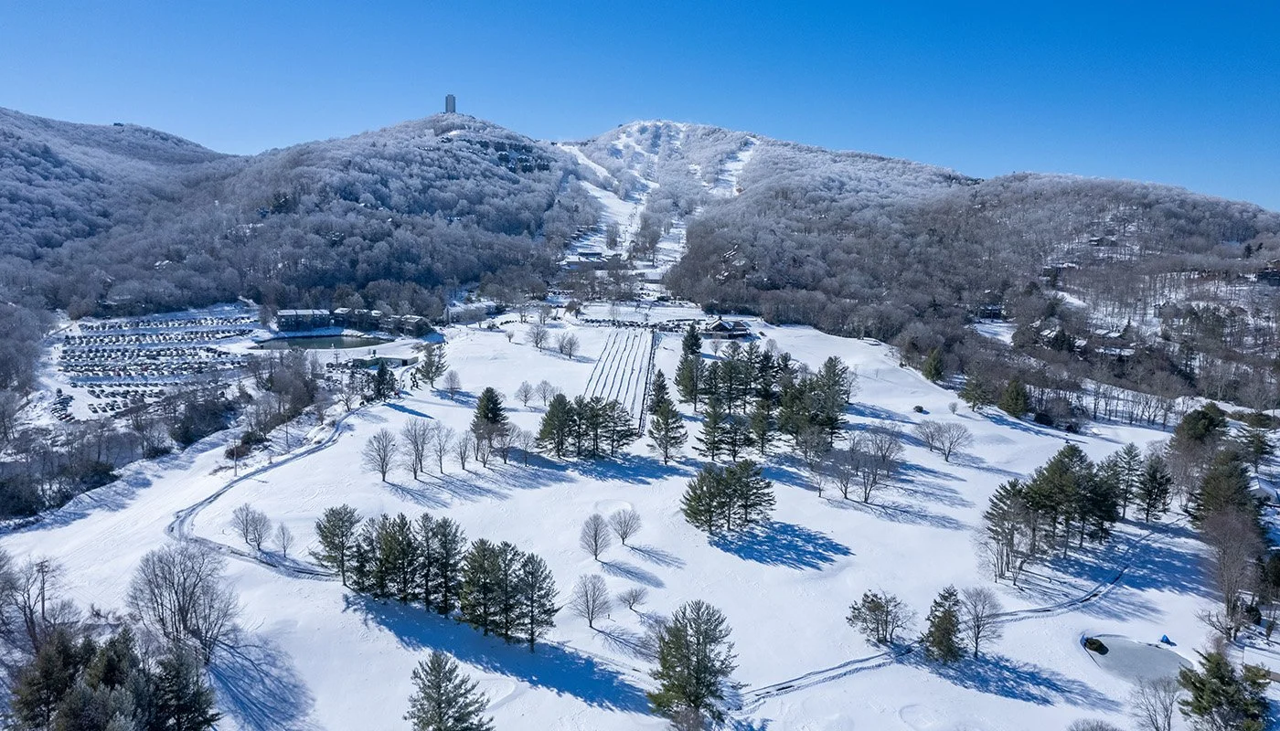 Sugar Mountain, North Carolina with a snow-covered landscape, trees, hills, and visible ski slopes on a clear, sunny day