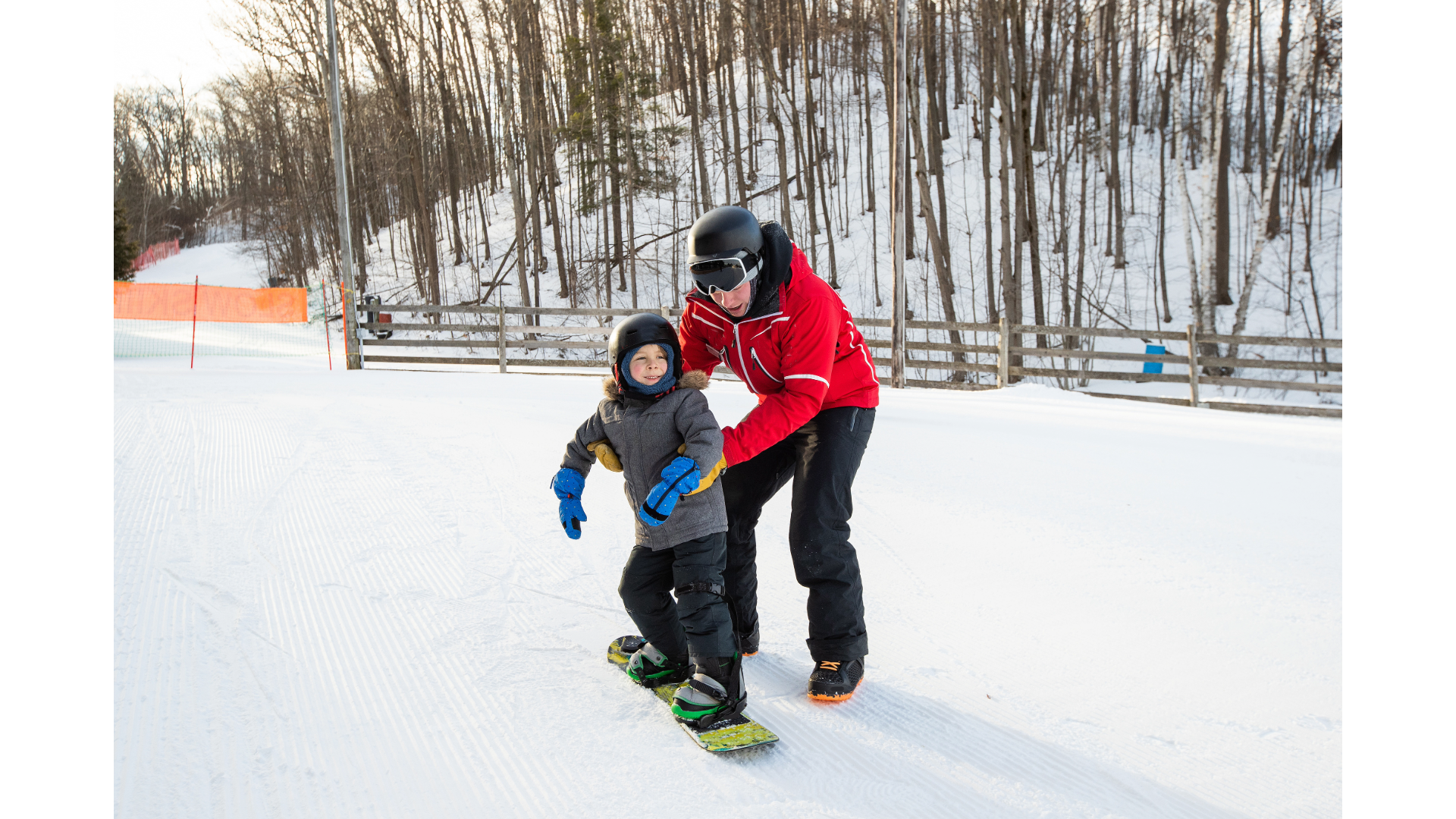 An adult and a child on snowboards at a North Carolina ski resort, with the adult teaching the child for the first time. They are both wearing helmets and winter gear, with a snowy wooded background.