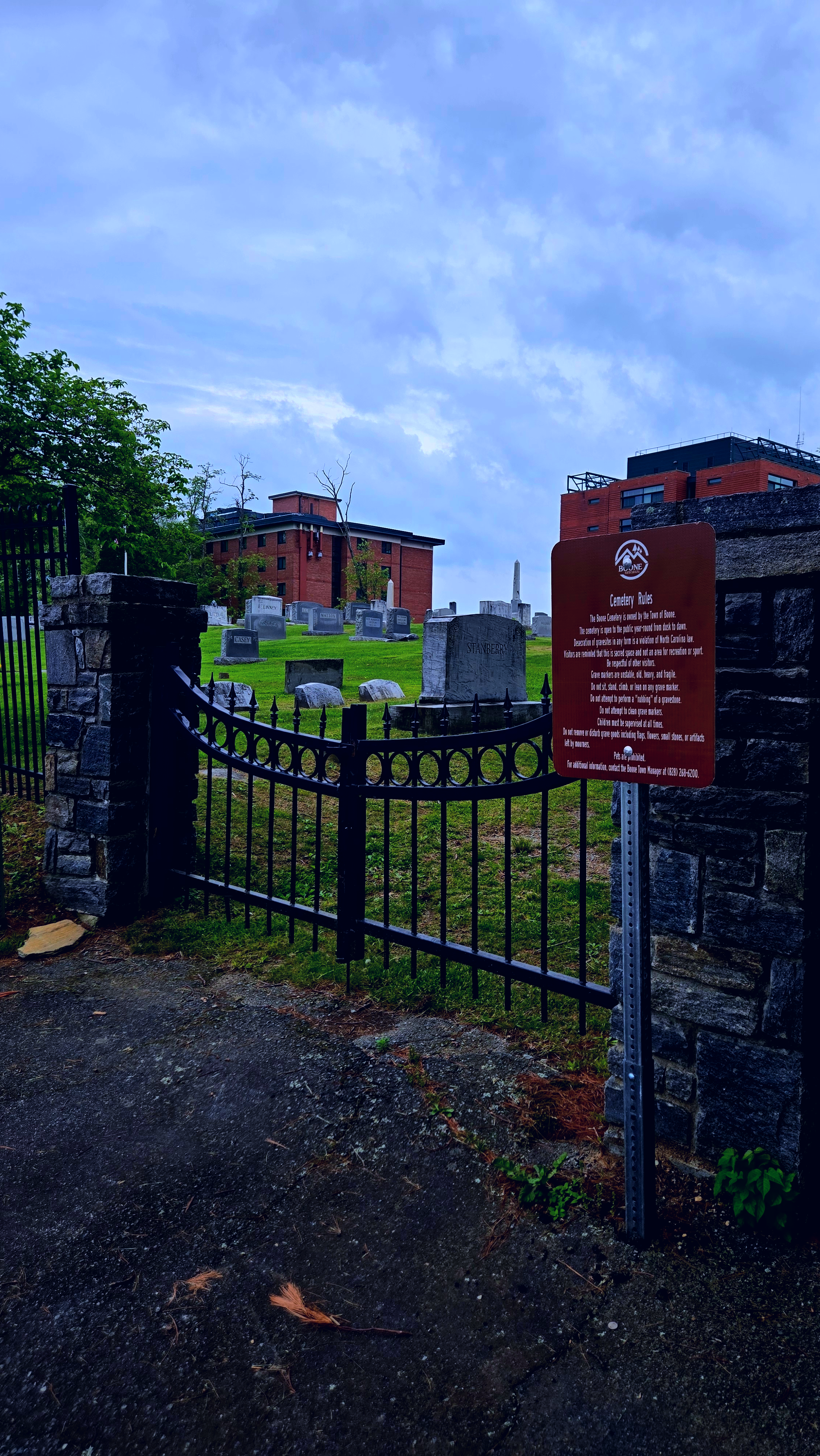 Cemetery entrance with metal fence and stone pillars, grave markers visible, red brick building and cloudy sky in background, sign with cemetery rules attached to fence post.