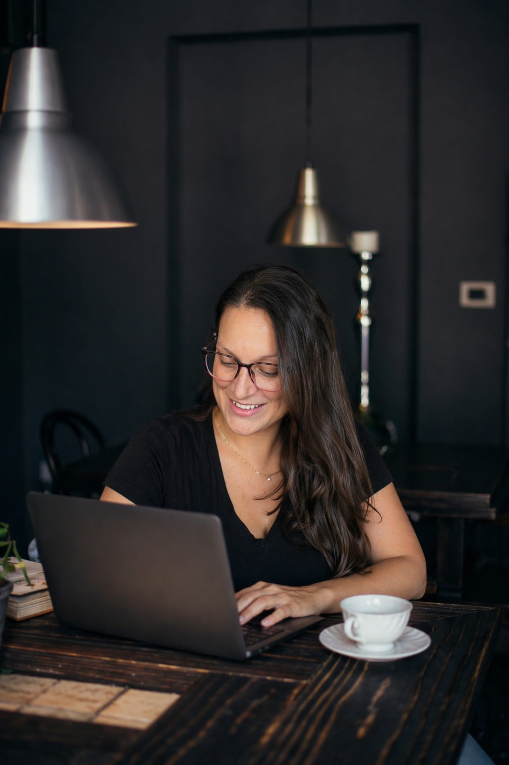 Owner of RivDesigns - A woman with long dark hair and glasses working on a laptop at a wooden table in a dark-colored room, with a cup on a saucer nearby and modern hanging lamps in the background.