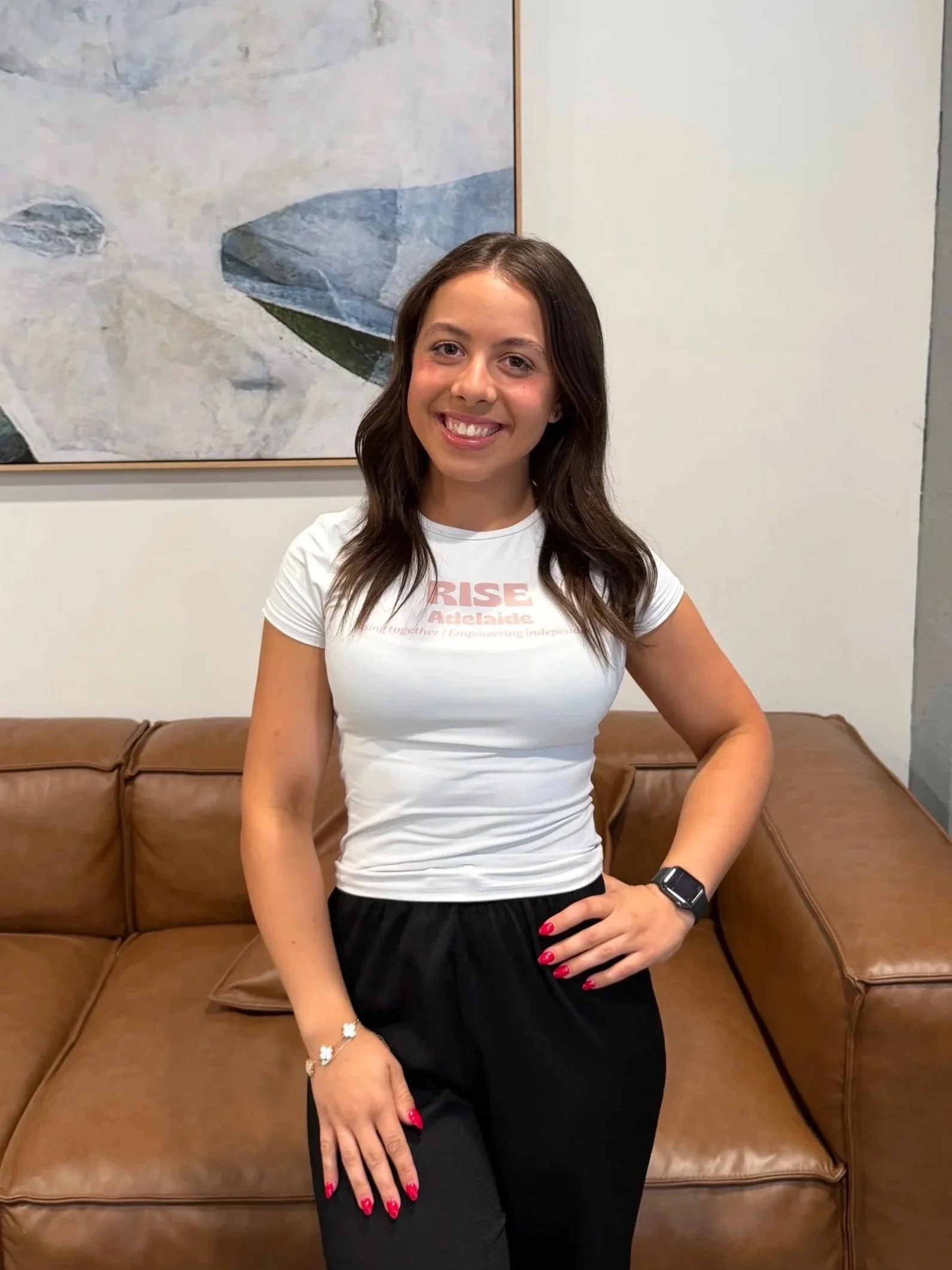Young woman with brown hair smiling and standing with her hand on her hip in a casual setting, wearing a white T-shirt, black pants, a watch, a bracelet, and red nail polish, sitting on a brown leather couch with artwork on the wall behind her.