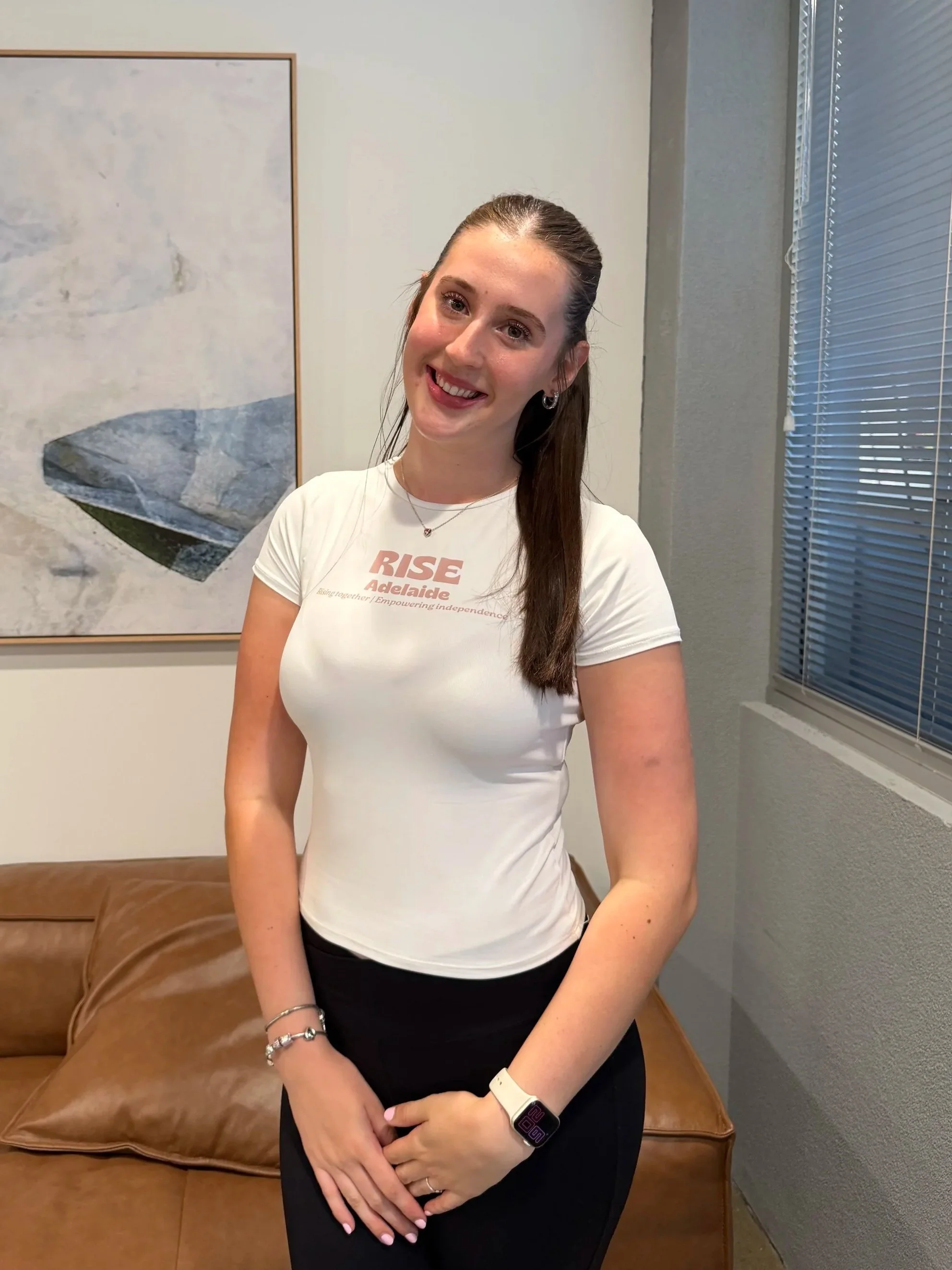 A young woman with long brown hair smiling, wearing a white t-shirt with 'RISE Adelaide' printed on it, black pants, an Apple Watch, and bracelets, standing in an office-like room with a brown couch, wall art, and blinds covering the window.