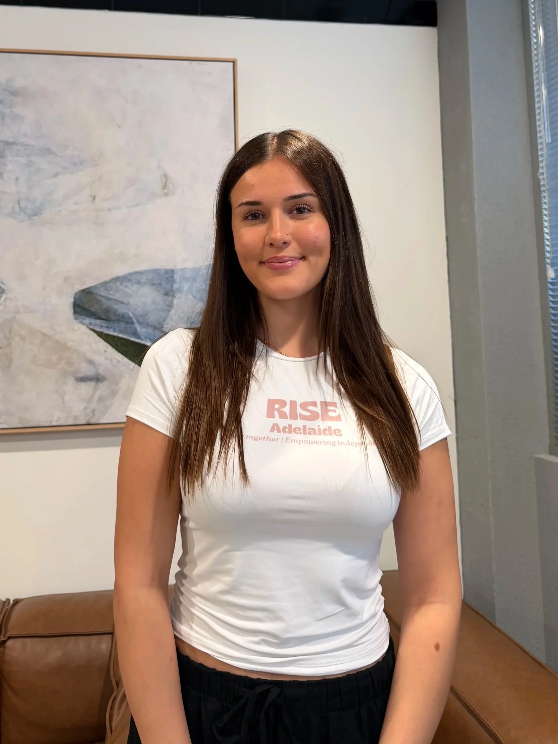 A young woman with long brown hair and fair skin, smiling, standing in a room with a painting and window behind her. She is wearing a white T-shirt with pink text and black pants.