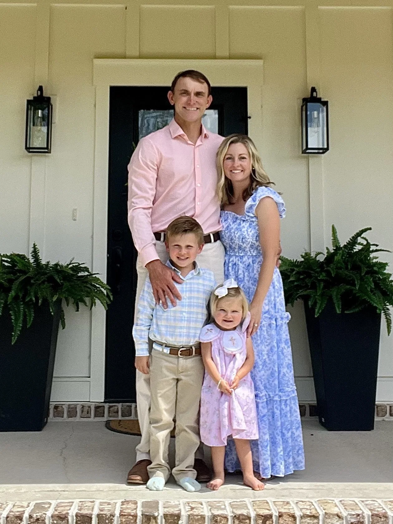 A family of five standing on a porch in front of a door, with two large potted plants on either side. The father is tall and wearing a pink shirt, the mother is wearing a blue dress, a boy is in a plaid shirt, and two young girls are dressed in light-colored dresses.