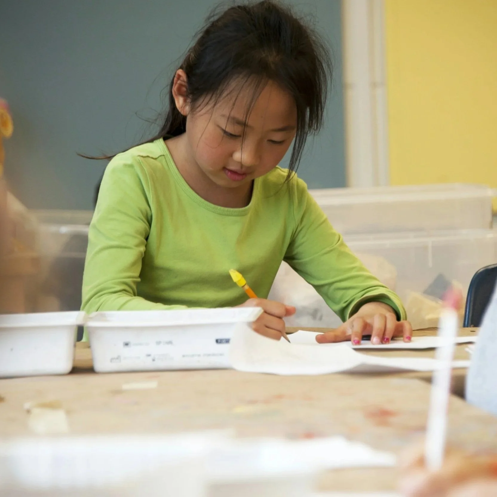 A young girl with dark hair wearing a bright green long-sleeve shirt is sitting at a table, writing on paper with a yellow pen, in a classroom or art room setting.