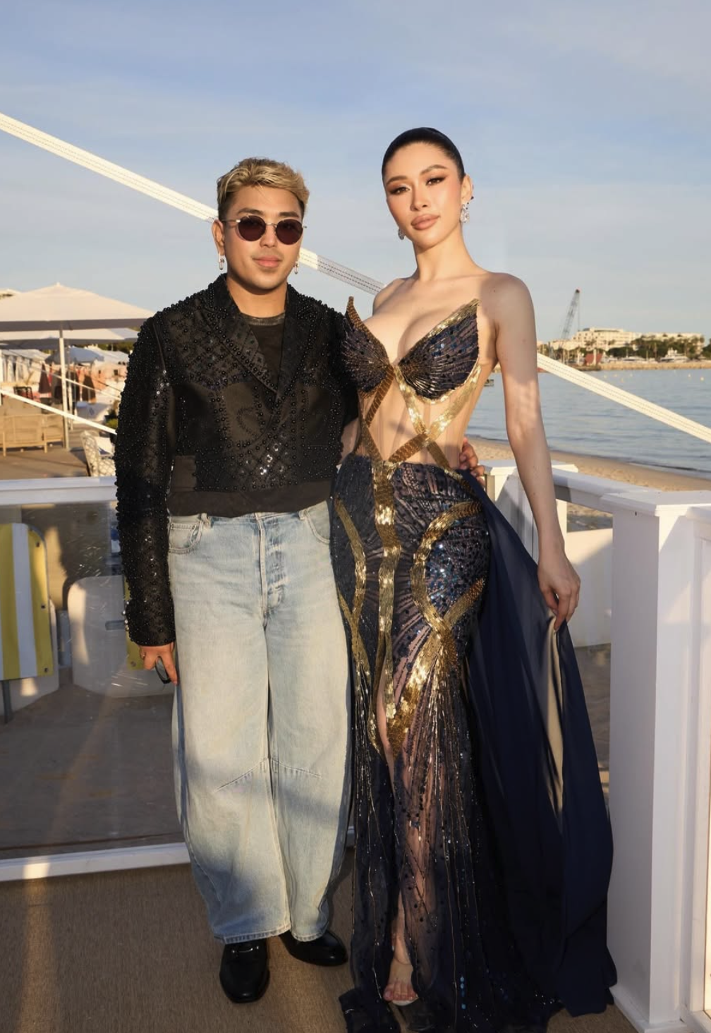 Two people standing together on a boat deck, with the water and a cityscape in the background during sunset. One person is dressed in a glamorous, gold and dark blue gown with intricate detail, and the other is wearing casual jeans and a black, glittery jacket with sunglasses.