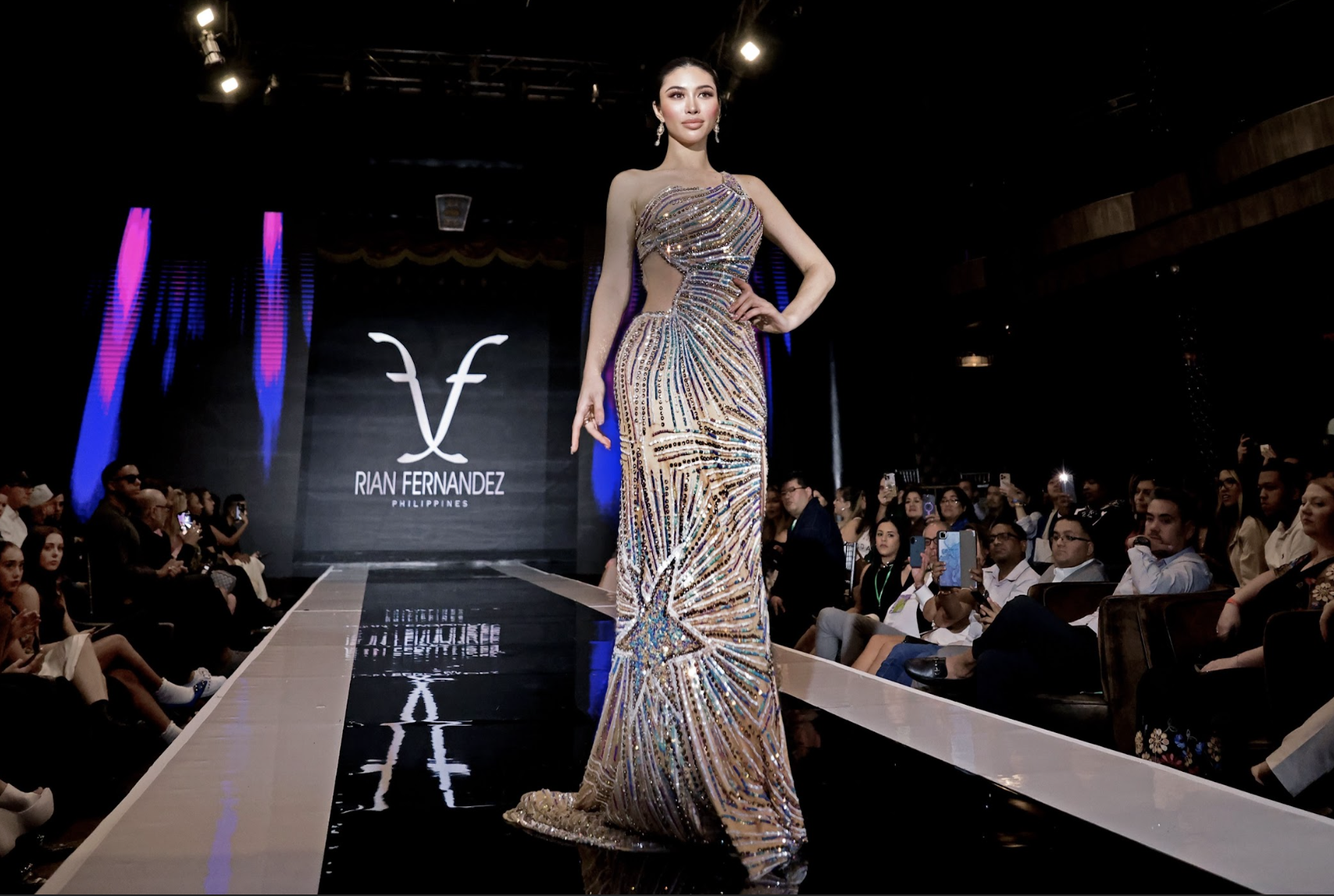 A woman walking on the runway at a fashion show, wearing a jeweled gown with intricate multicolored patterns and cutouts, with the name 'Rian Fernandez Philippines' displayed on a screen in the background.