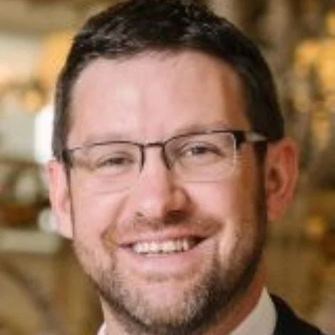 A smiling man with glasses and facial hair, wearing a suit and tie, in an indoor setting with blurred background.