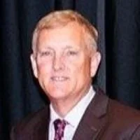 A man in a dark suit and tie standing in front of a dark curtain.
