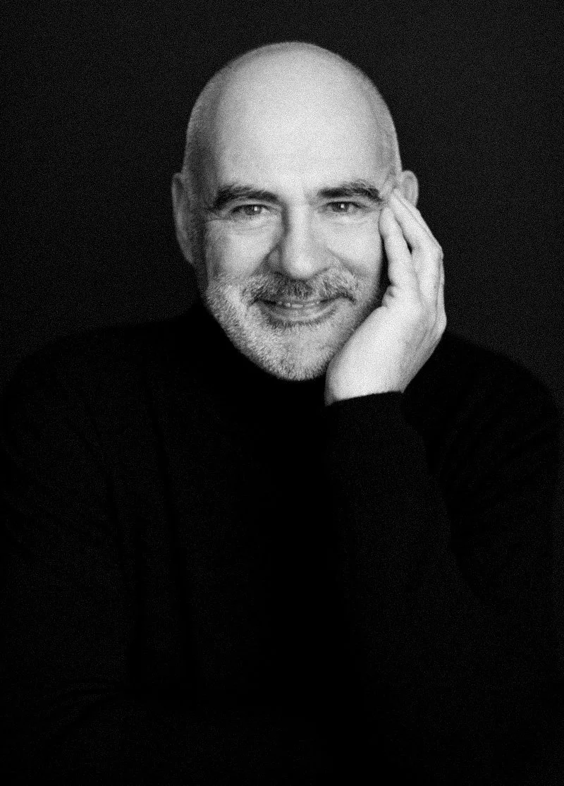 Black and white portrait of a smiling bald man with a beard, resting his face on his hand, wearing a dark sweater, against a dark background.