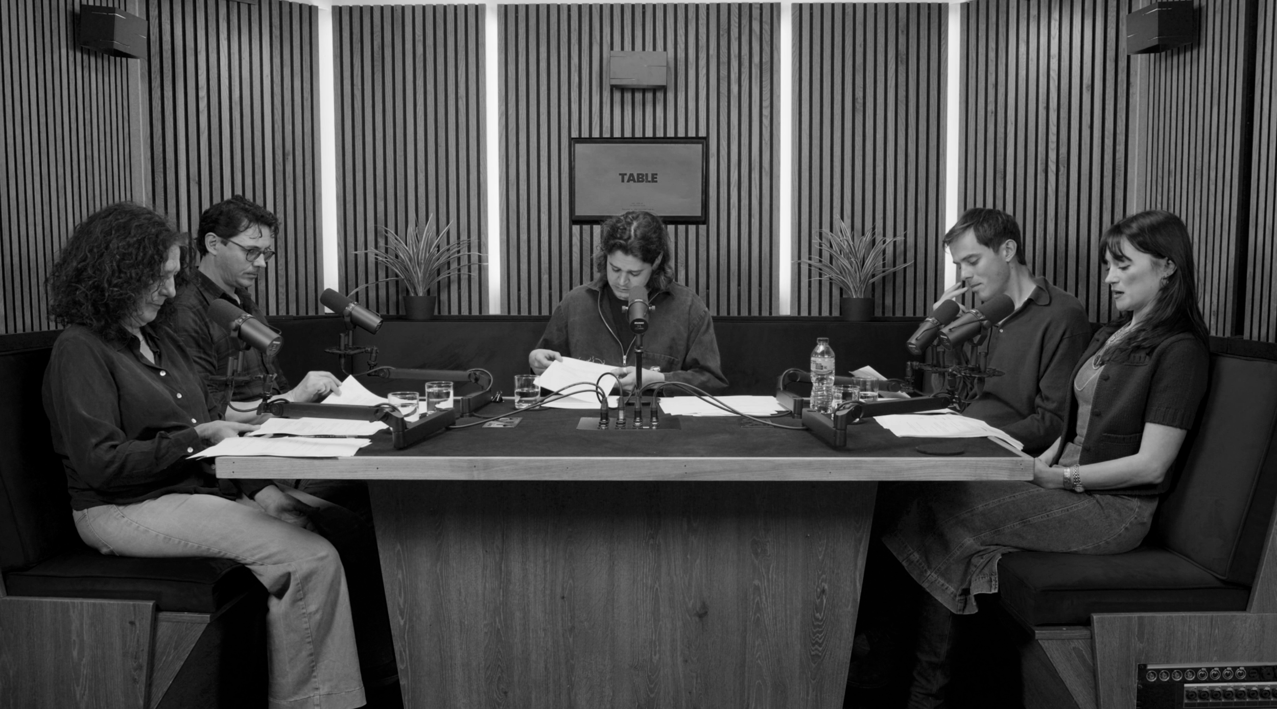 Six people sit around a rectangular table in a recording studio, with microphones and papers in front of them, engaged in a discussion.
