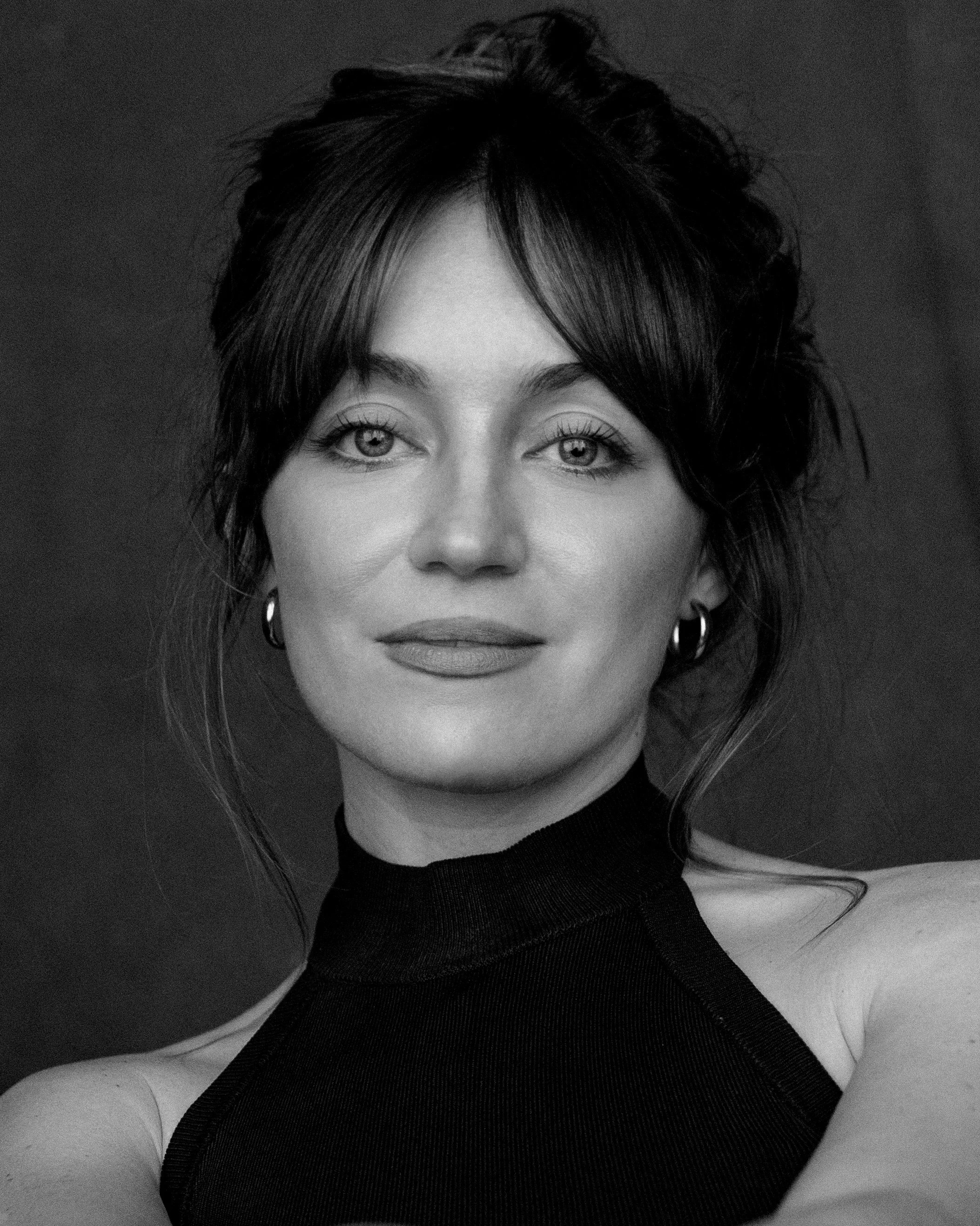 Black and white close-up photo of a woman with light-colored eyes, dark wavy hair, and hoop earrings, wearing a black sleeveless top.