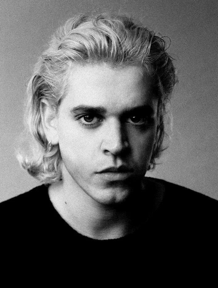 Black and white portrait of a young man with light-colored, wavy hair and intense stare, wearing a black shirt.