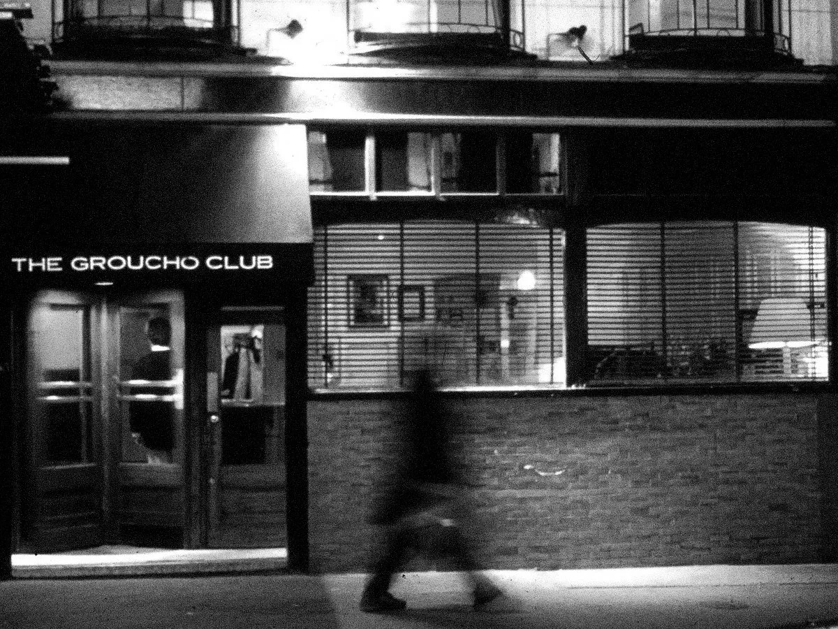 Nighttime view of The Groucho Club entrance with a person walking past on the sidewalk, black and white photo.