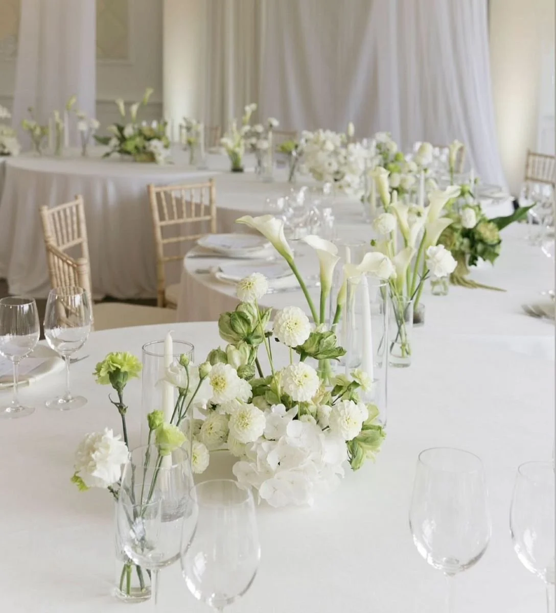 Elegant wedding reception table with white floral centerpieces, glassware, and gold chairs in a bright room with cream curtains.