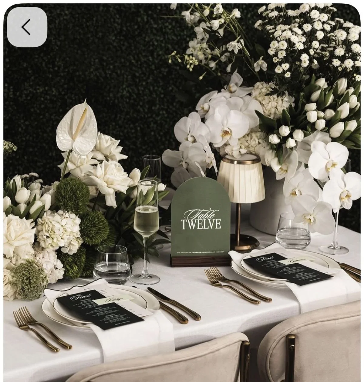 Elegant wedding reception table decorated with white flowers, including orchids, tulips, and anthuriums, arranged with greenery. The table has gold cutlery, glassware, and a black menu. A green table sign reads 'Table Twelve' with a small lampshade in the background.