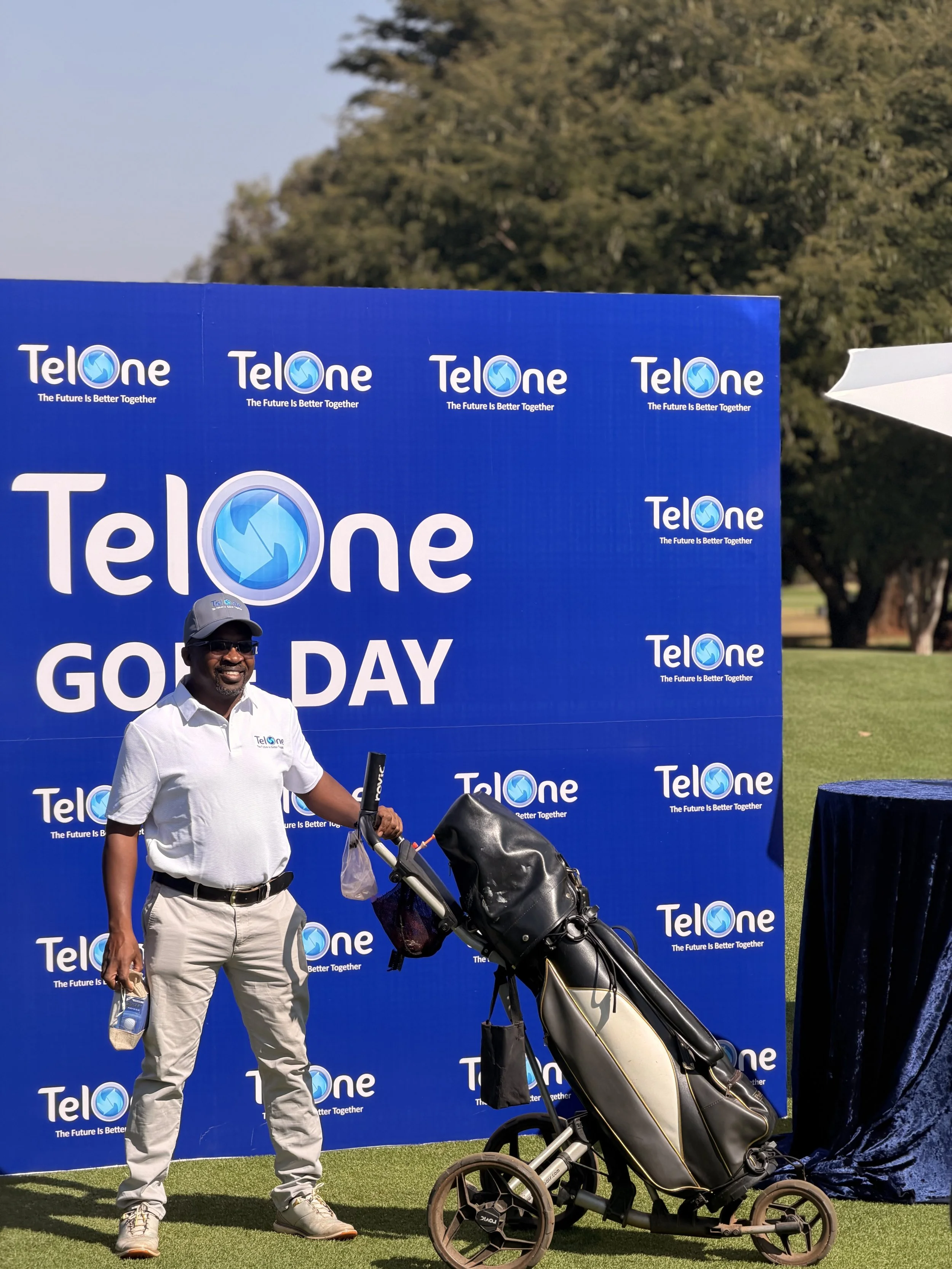 A man standing next to a golf trolley with golf clubs at a golf day event, in front of a blue backdrop with the TelOne logo and slogan.