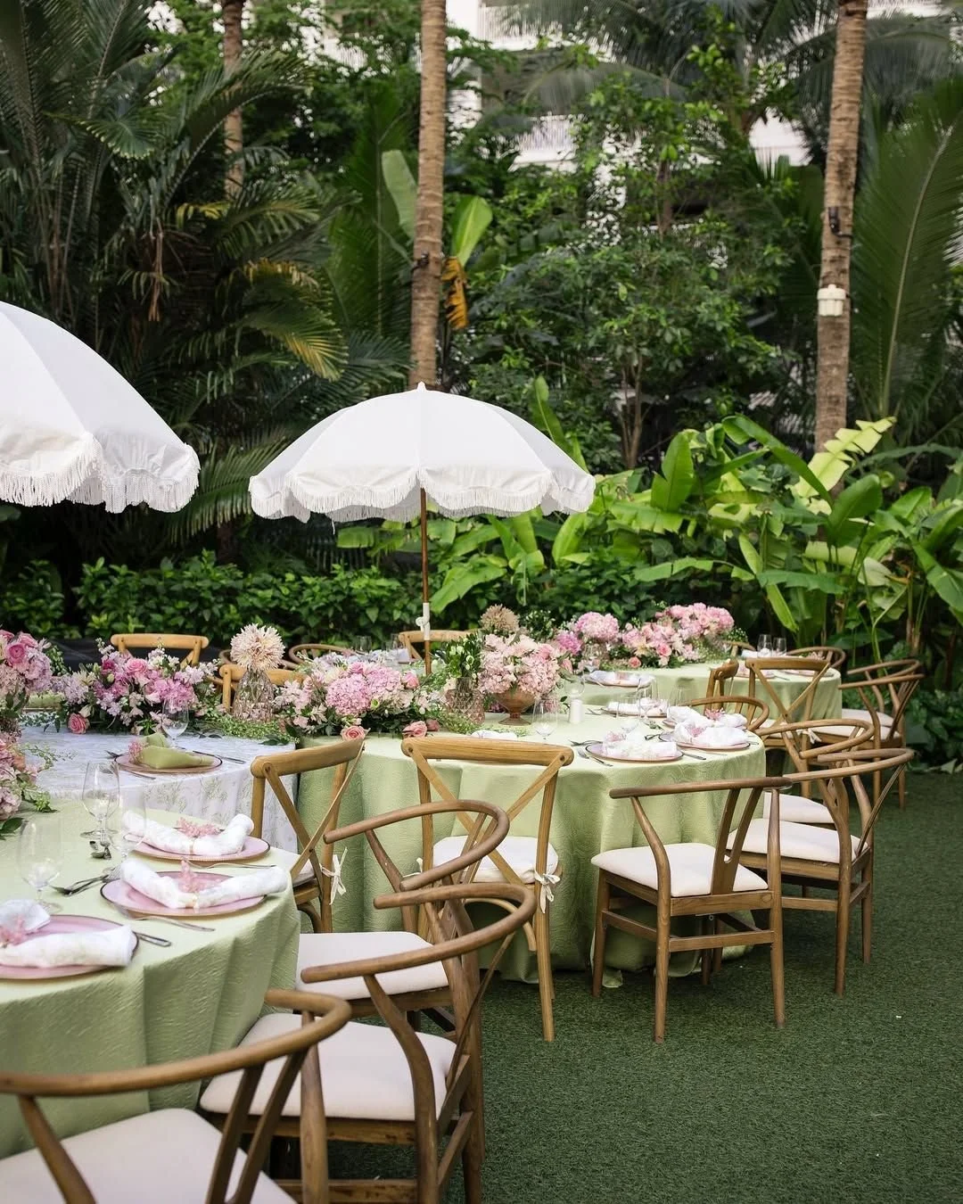Outdoor dining setup with round tables covered in green tablecloths, decorated with pink and white flower arrangements, set with plates, glasses, and napkins, shaded by white umbrellas, surrounded by lush green tropical plants and trees.