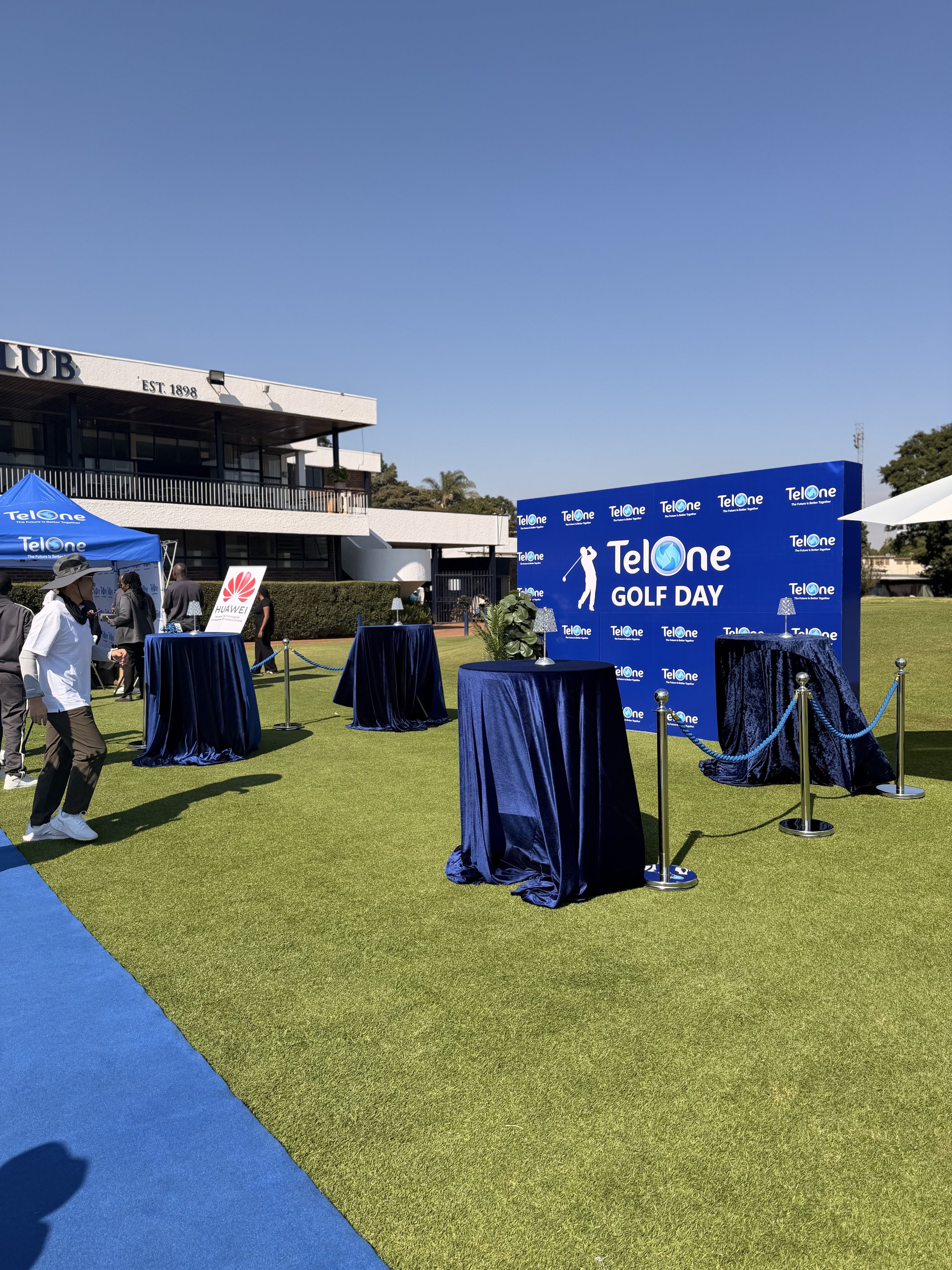 Outdoor event setup with blue tables, a large blue backdrop reading "TelOne GOLF DAY," and a few people walking on a green grass field. There is a building in the background.