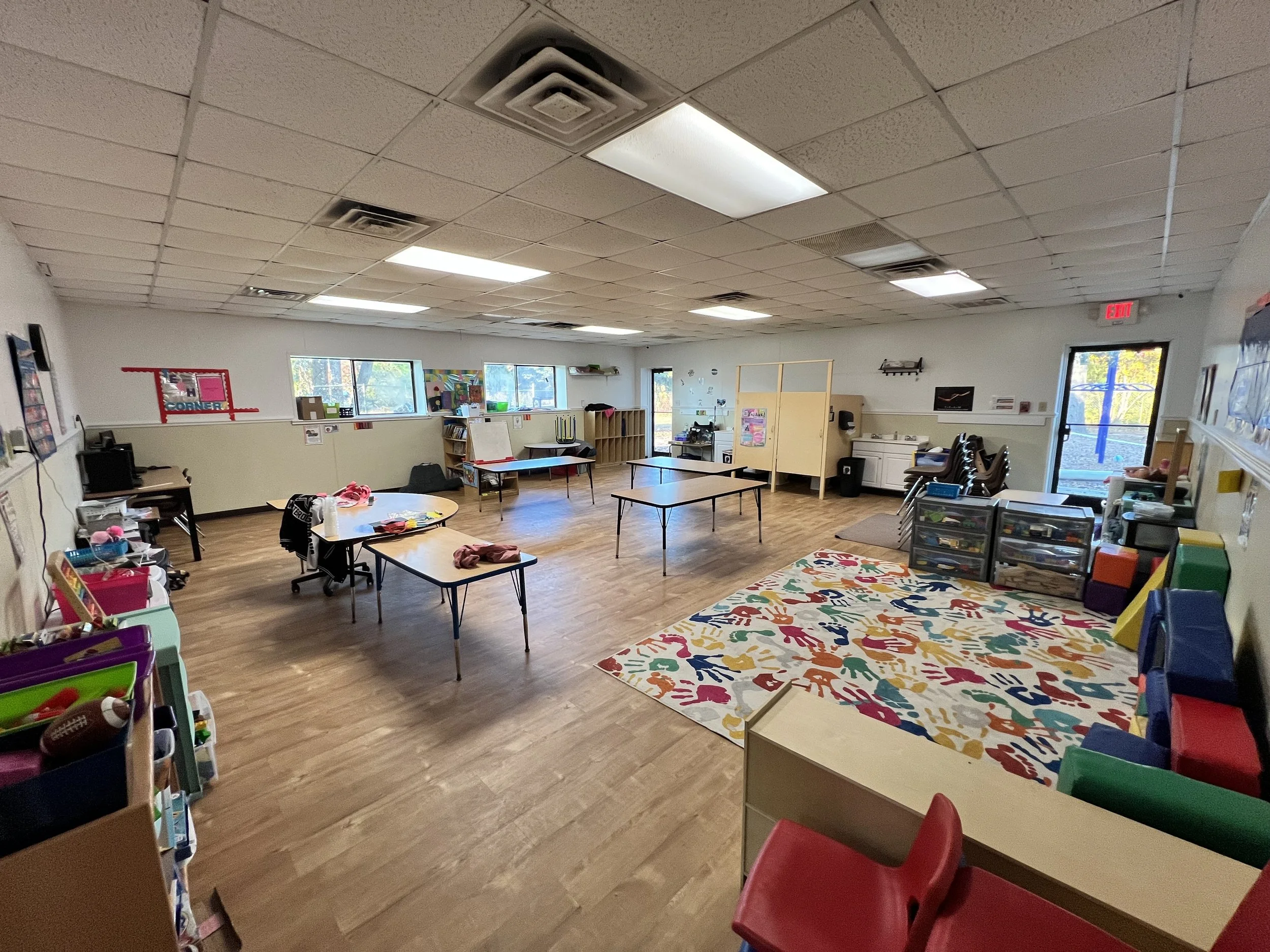A spacious classroom with tables and chairs, colorful storage bins, a patterned rug, windows allowing natural light, and educational decorations on the walls.