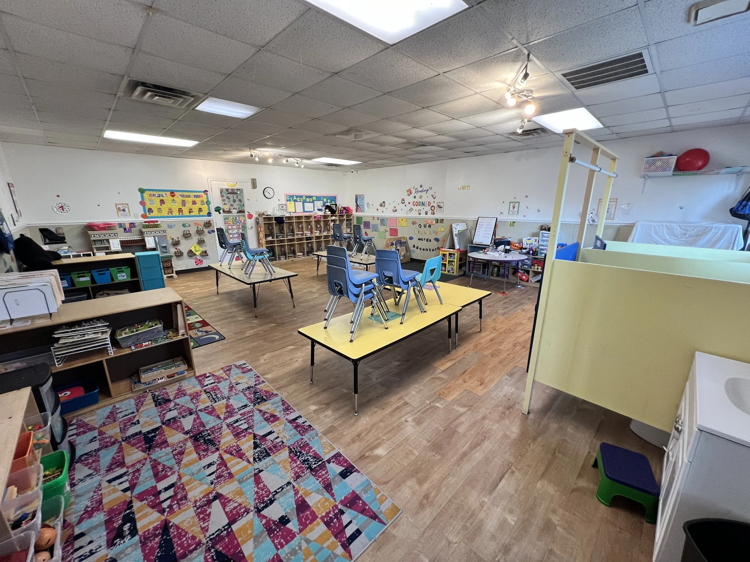 A classroom with small chairs stored on top of tables, colorful cubbies, educational posters, and toys, with a hardwood floor and a detailed rug near the front.