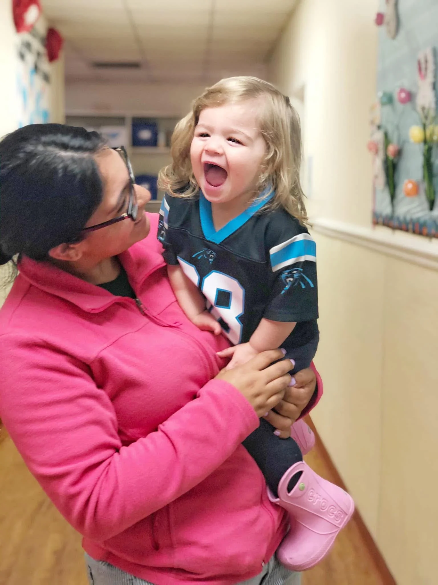 A woman in a pink jacket holding a young girl with curly hair, who is laughing and wearing a sports jersey and pink shoes, in a hallway with colorful decorations on the wall.